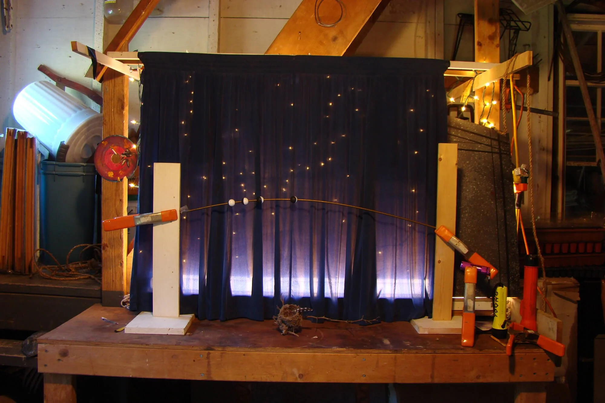 A puppet theater setup with a dark curtain backdrop and string lights, mounted on a wooden platform in a workshop or garage, with tools and materials around.