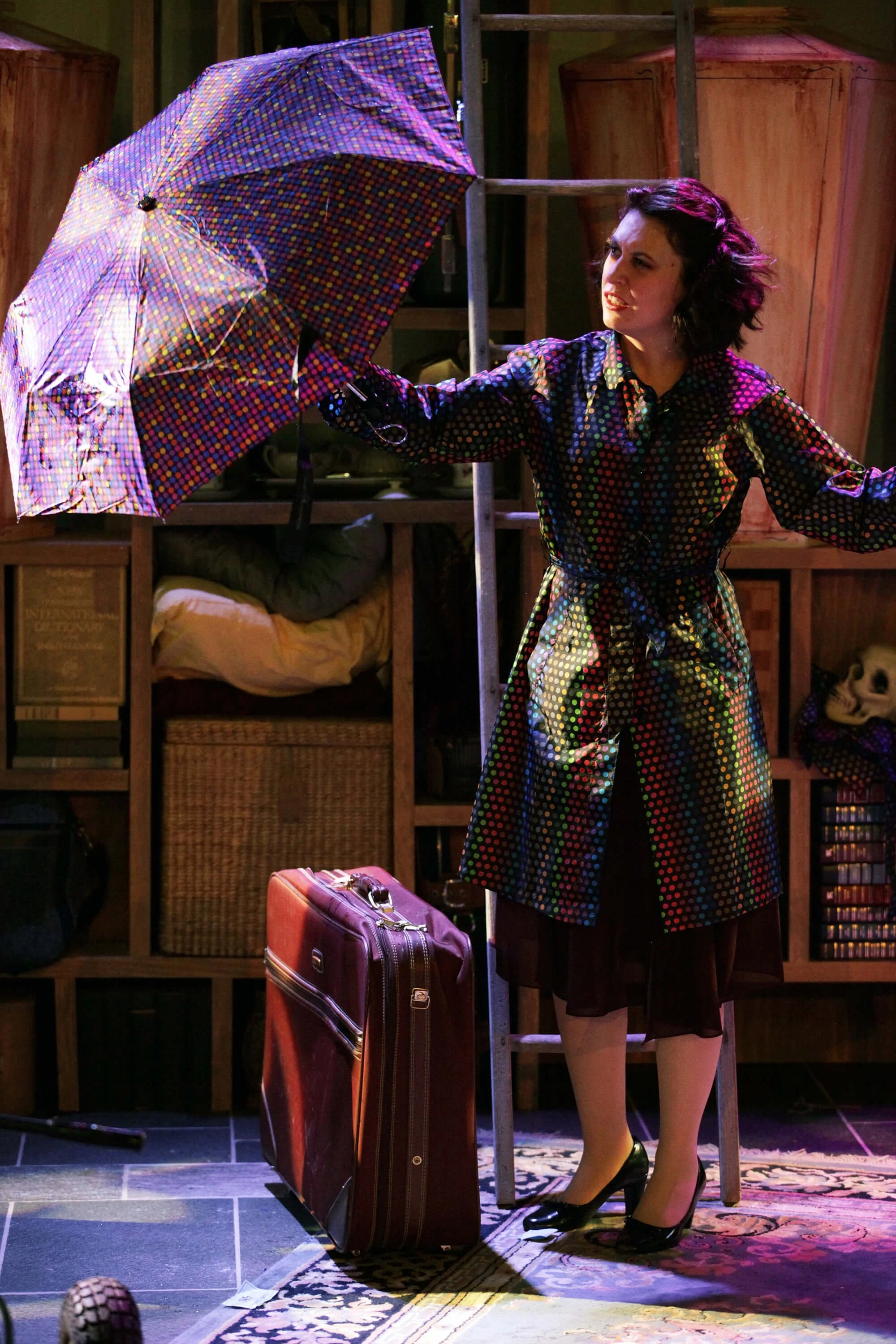 Woman with curly dark hair wearing a colorful, patterned coat and black heels holding an umbrella on a stage set, with a suitcase in front and a wooden shelving unit behind her.