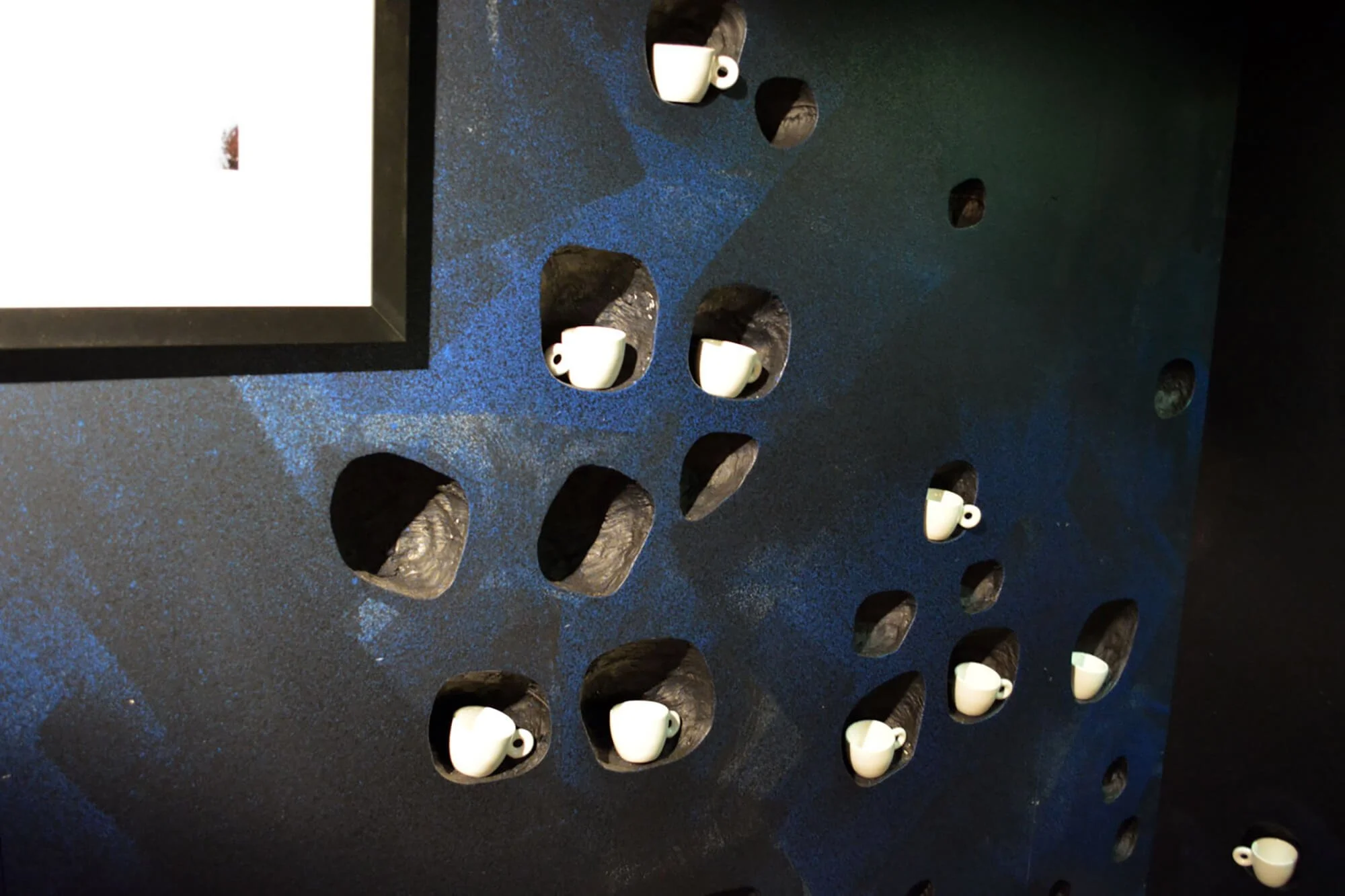 Display with small white coffee cup ornaments placed in cave-like holes on a blue textured wall, with part of a white sign or photo frame visible in the upper left corner.
