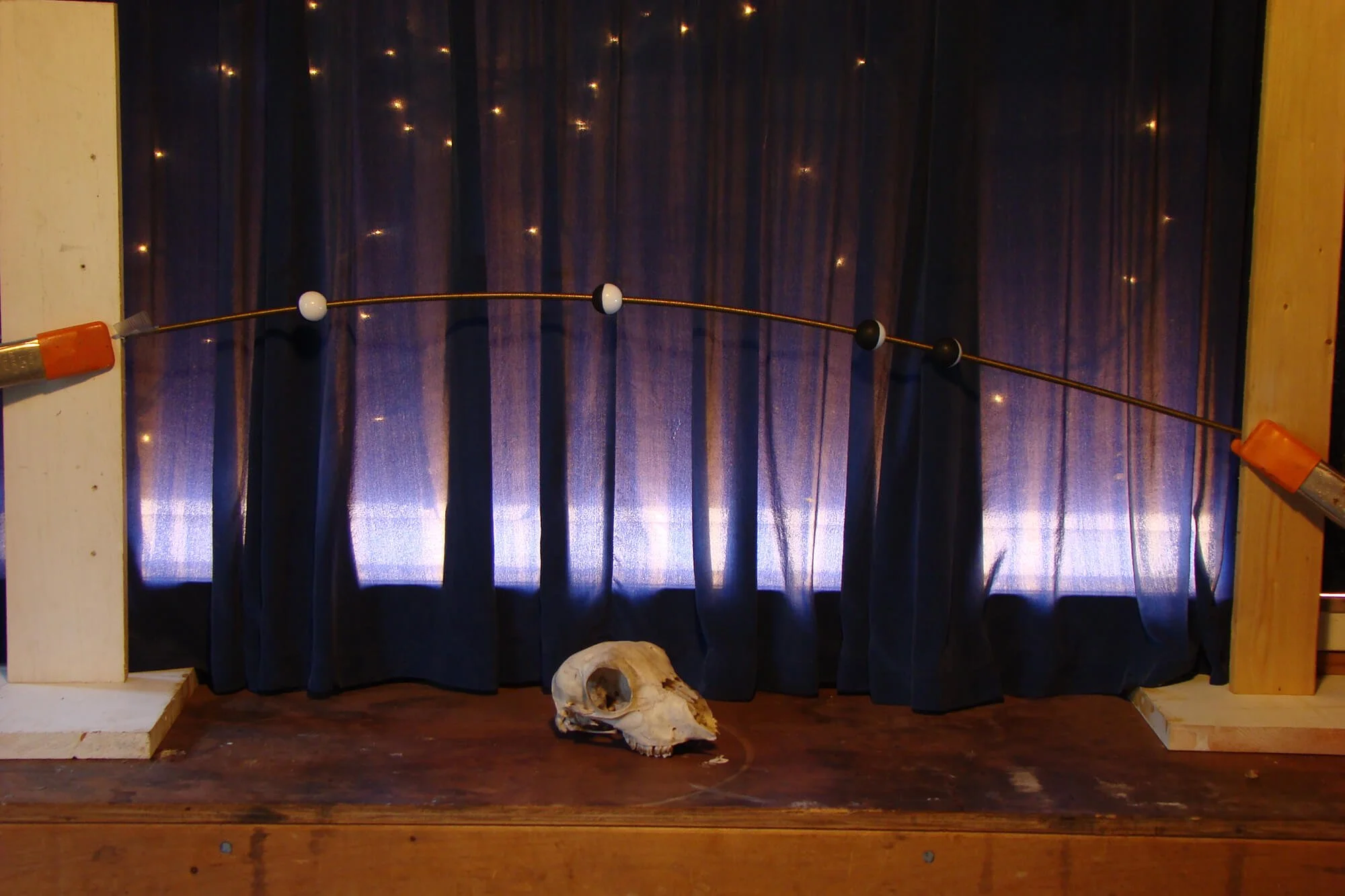 A view of a dark curtain with star-like light reflections, a wooden platform, a small animal skull on the floor, and a string of white and black beads with orange clips attached, suspended between two wooden supports.