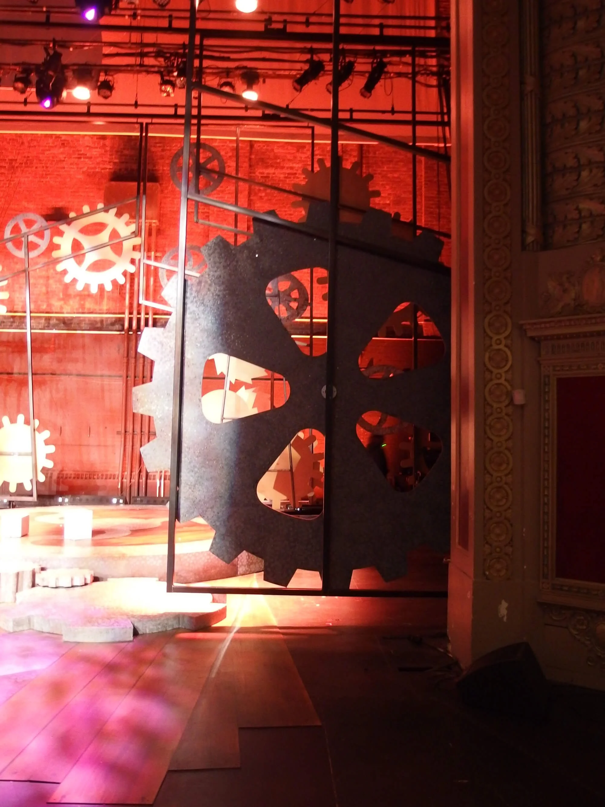 A large metal gear and cog mechanism with cutouts, suspended in a decorated room with stage lighting and a brick wall backdrop.