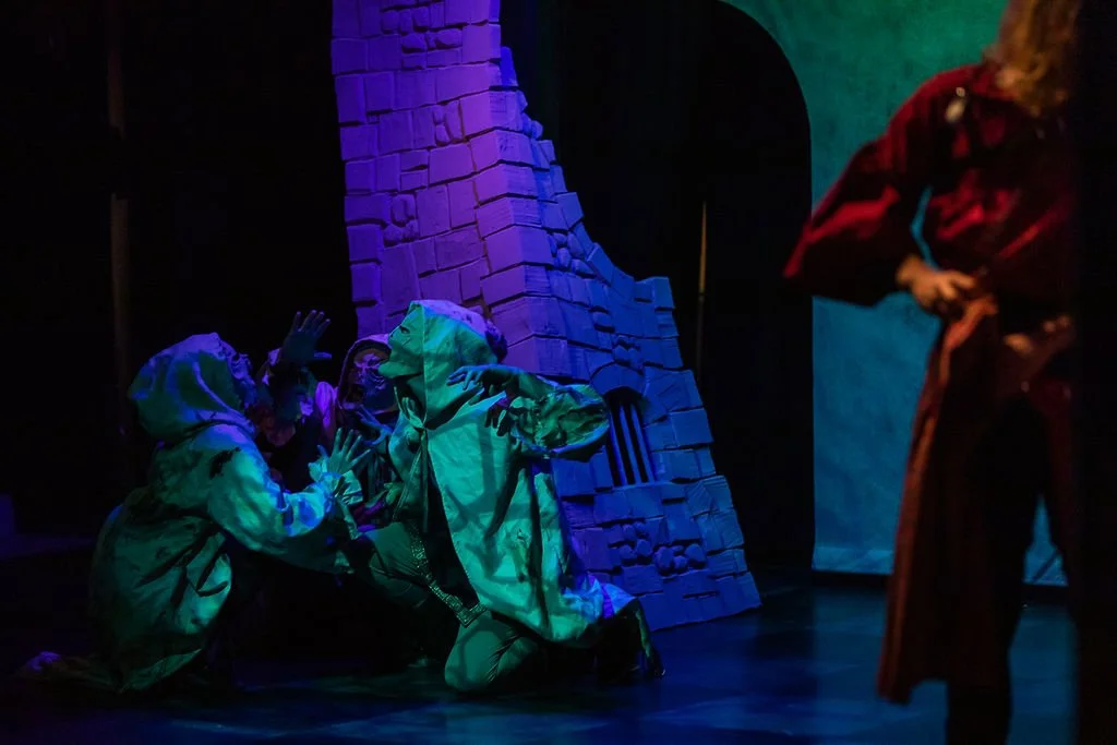 Three children wearing hooded jackets are on the stage, with one kneeling and the other two standing, appearing to act out a scene. A person dressed in a long, dark red cloak stands nearby. The background features a stone wall with blue, purple, and 