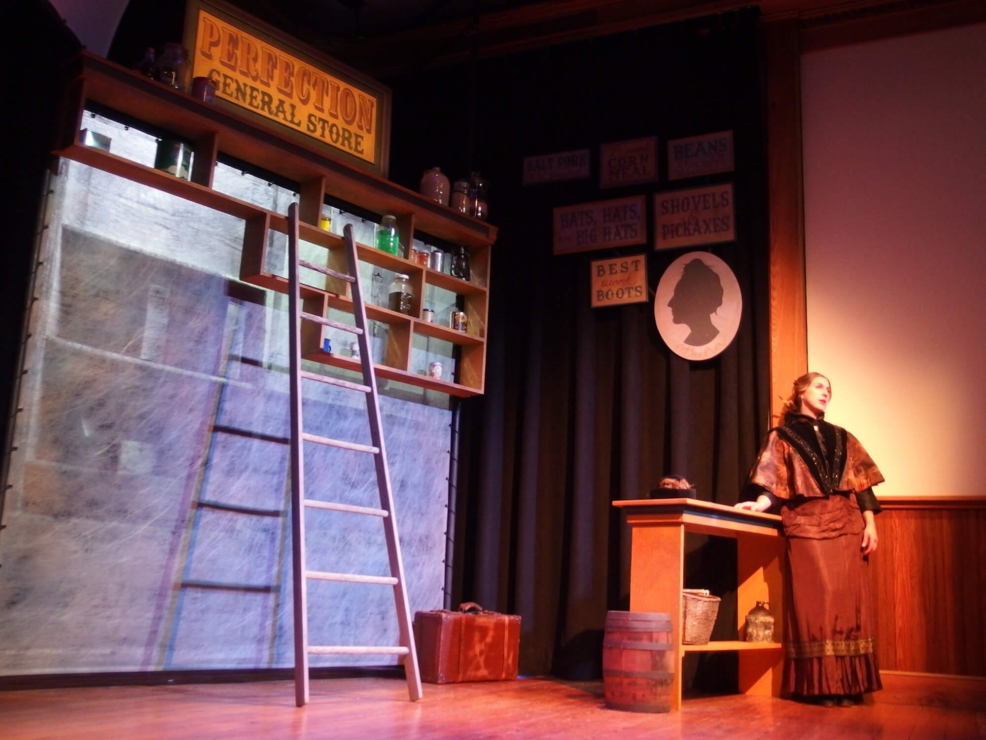 Stage set with a ladder, shelves with jars, vintage trunk, barrel, and a mannequin dressed in period clothing, with dark curtains and vintage signs on the wall.