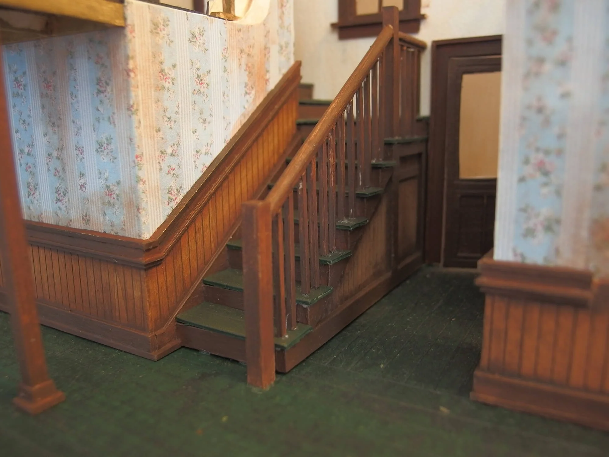 Model of an indoor staircase with wooden railing and floral wallpaper on the wall.