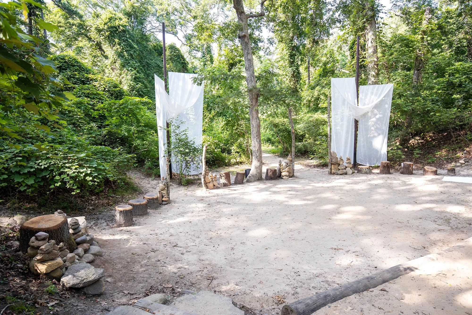 An outdoor natural setting with a cleared dirt area surrounded by lush green trees and bushes. There are white fabric panels hanging between dark poles, rocks, and tree stumps arranged to form a seating or border area, suggesting it is set up for an 