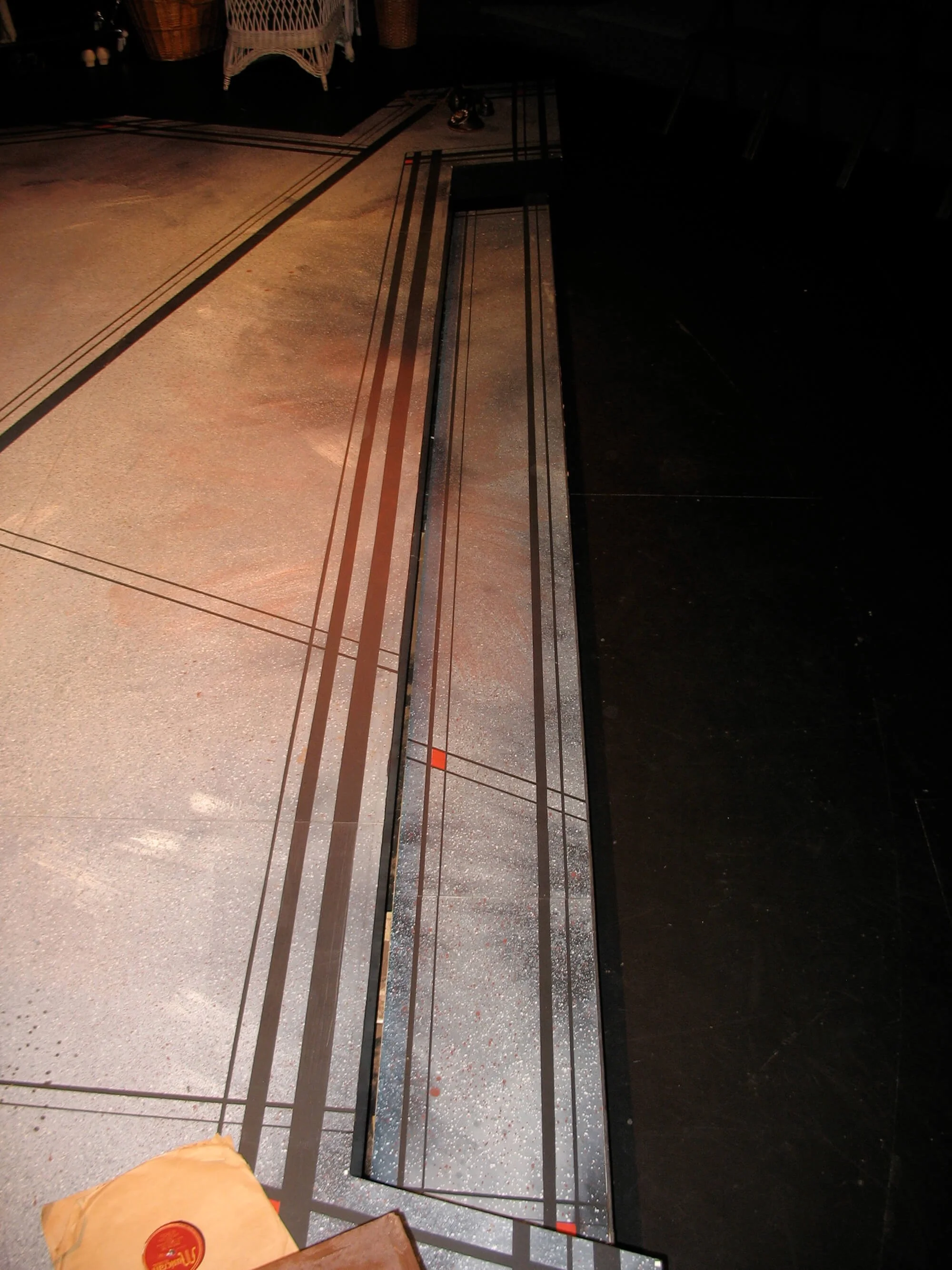 Close-up of a geometric tile pattern on a floor, featuring a long, rectangular metallic element with thin black lines and small red accents, with a dark background to the side.