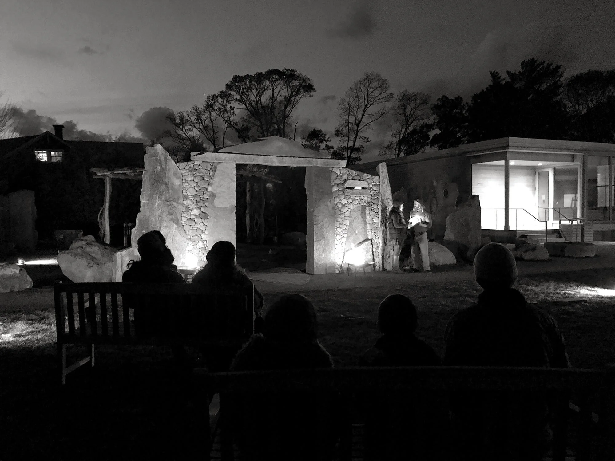 Nighttime outdoor scene with a small audience sitting on benches, watching two performers illuminated by a spotlight, in front of a stone arch and modern building.