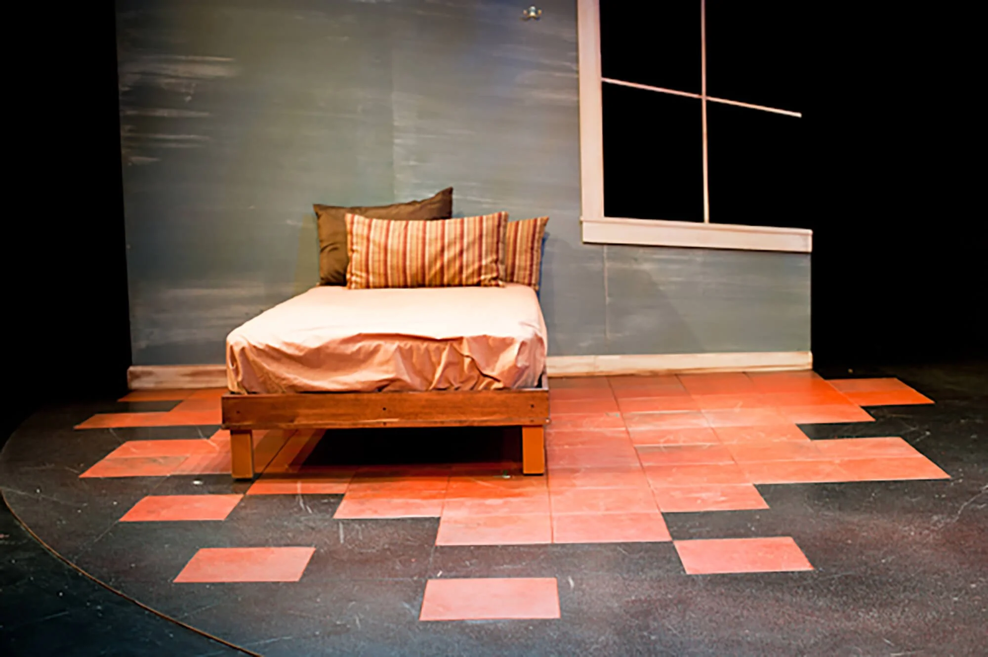 A small wooden bed with a white sheet and three pillows, two with striped patterns and one plain dark, set against a blue wall with a window. The floor has a pattern of black and pinkish-orange tiles.