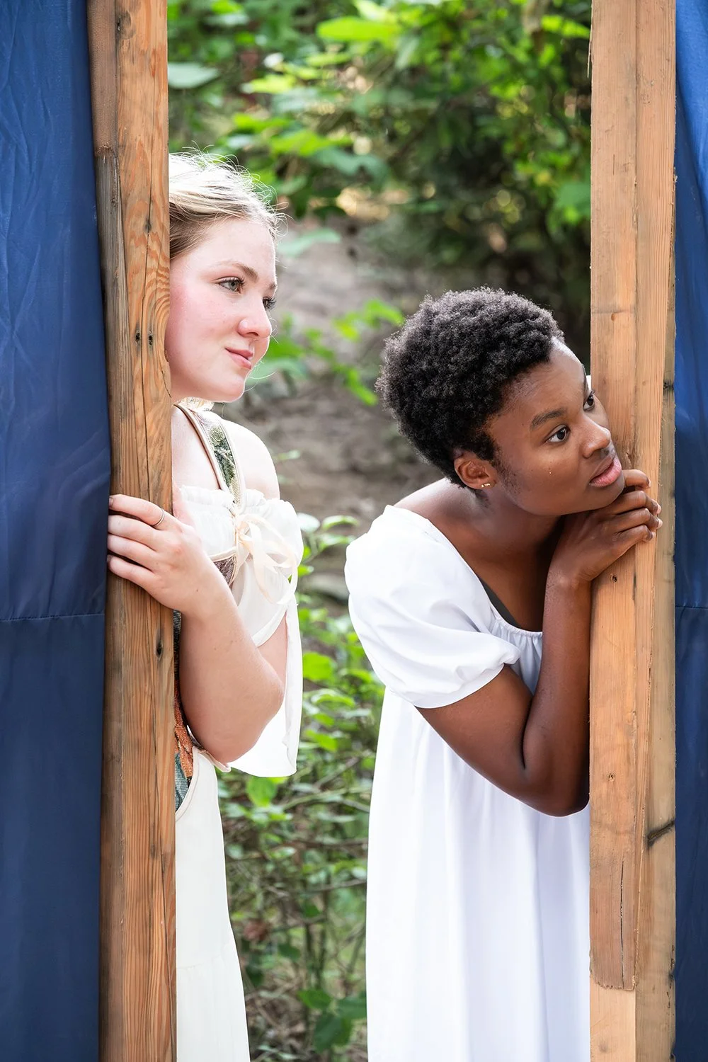 Two women peering through wooden frames with blue fabric, in an outdoor setting with green foliage.
