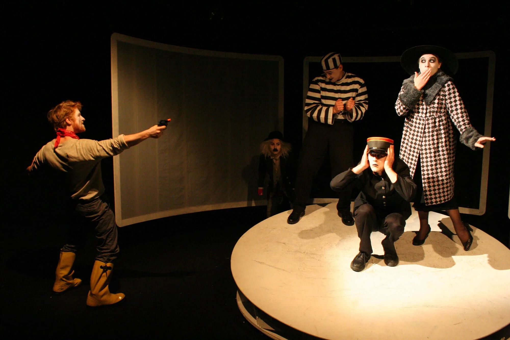 A theatrical performance with five actors in costume on a stage with a black background. One actor is pointing a prop gun at another actor dressed in dark clothing and a hat, squatting with a paper hat. Two other actors are standing, one in a striped