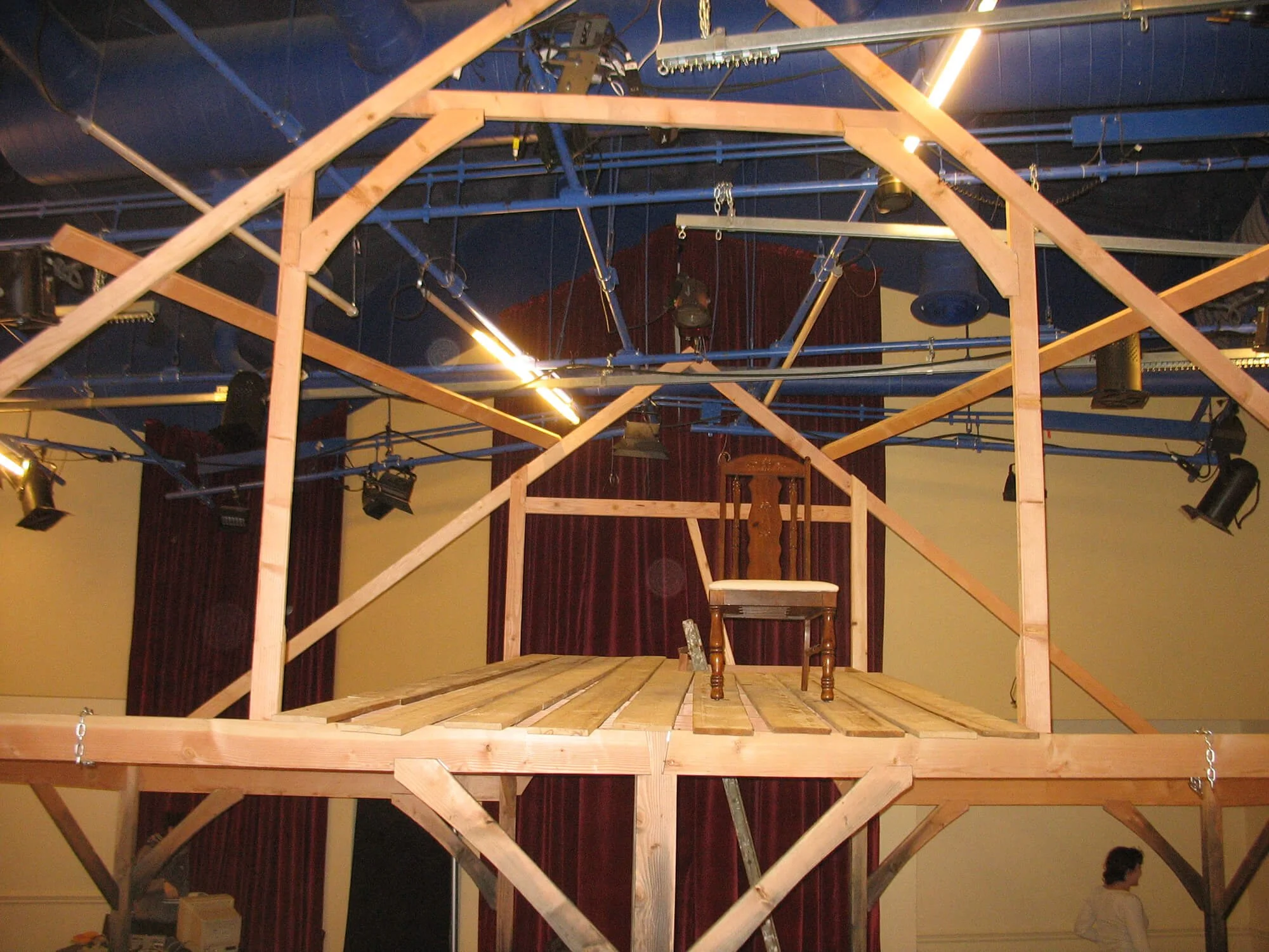 A wooden stage with a small chair on top, under construction, in a room with stage lighting and red curtains in the background.
