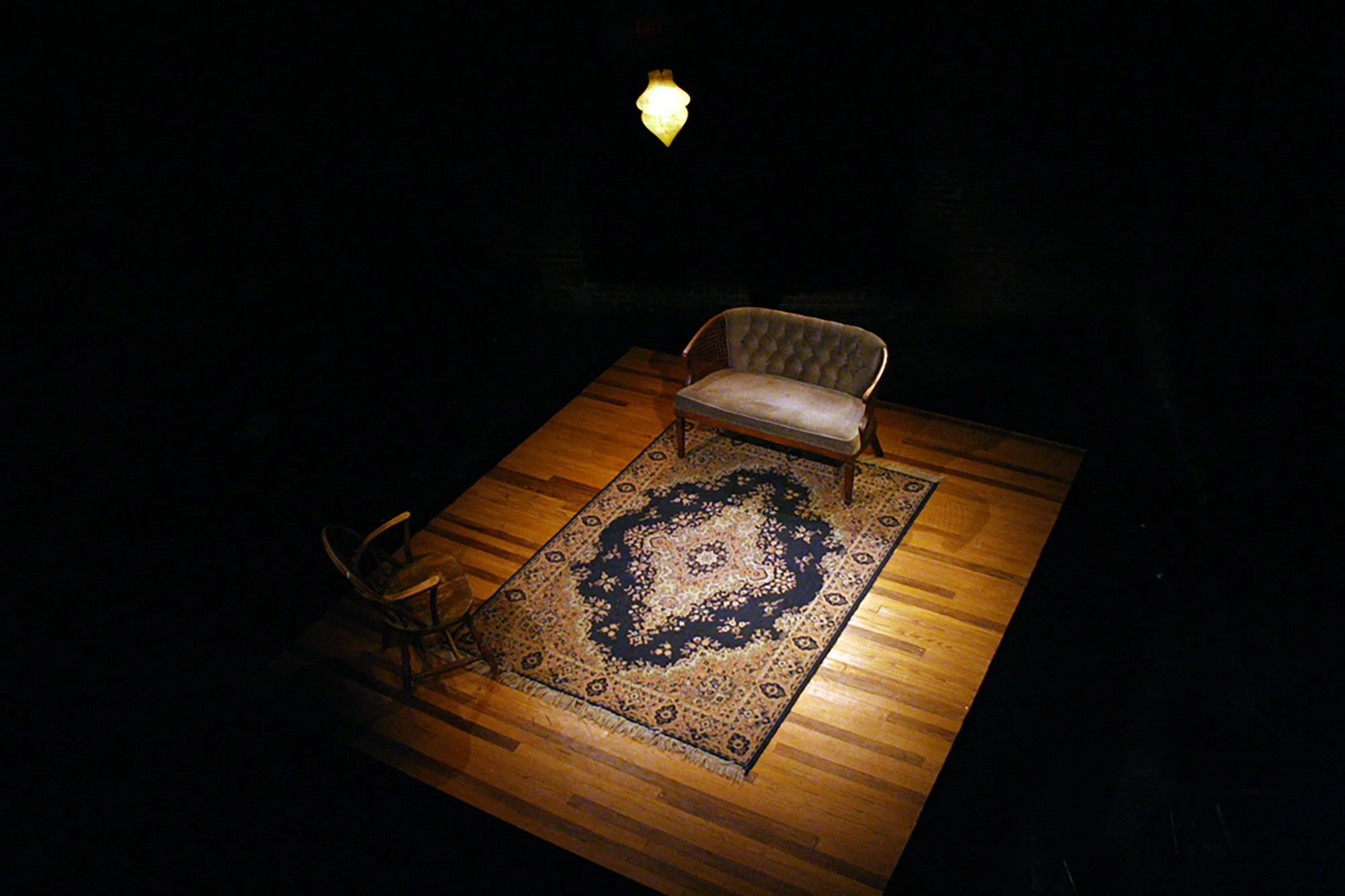 A vintage living room setup with a sofa, a wooden rocking chair, a patterned area rug, and a hanging ceiling lamp in a dark space.
