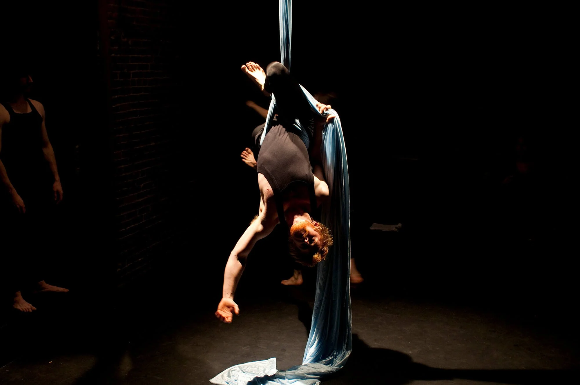 A female aerial silk performer hanging upside down in a dark room with a brick wall, with others standing nearby in the shadows.