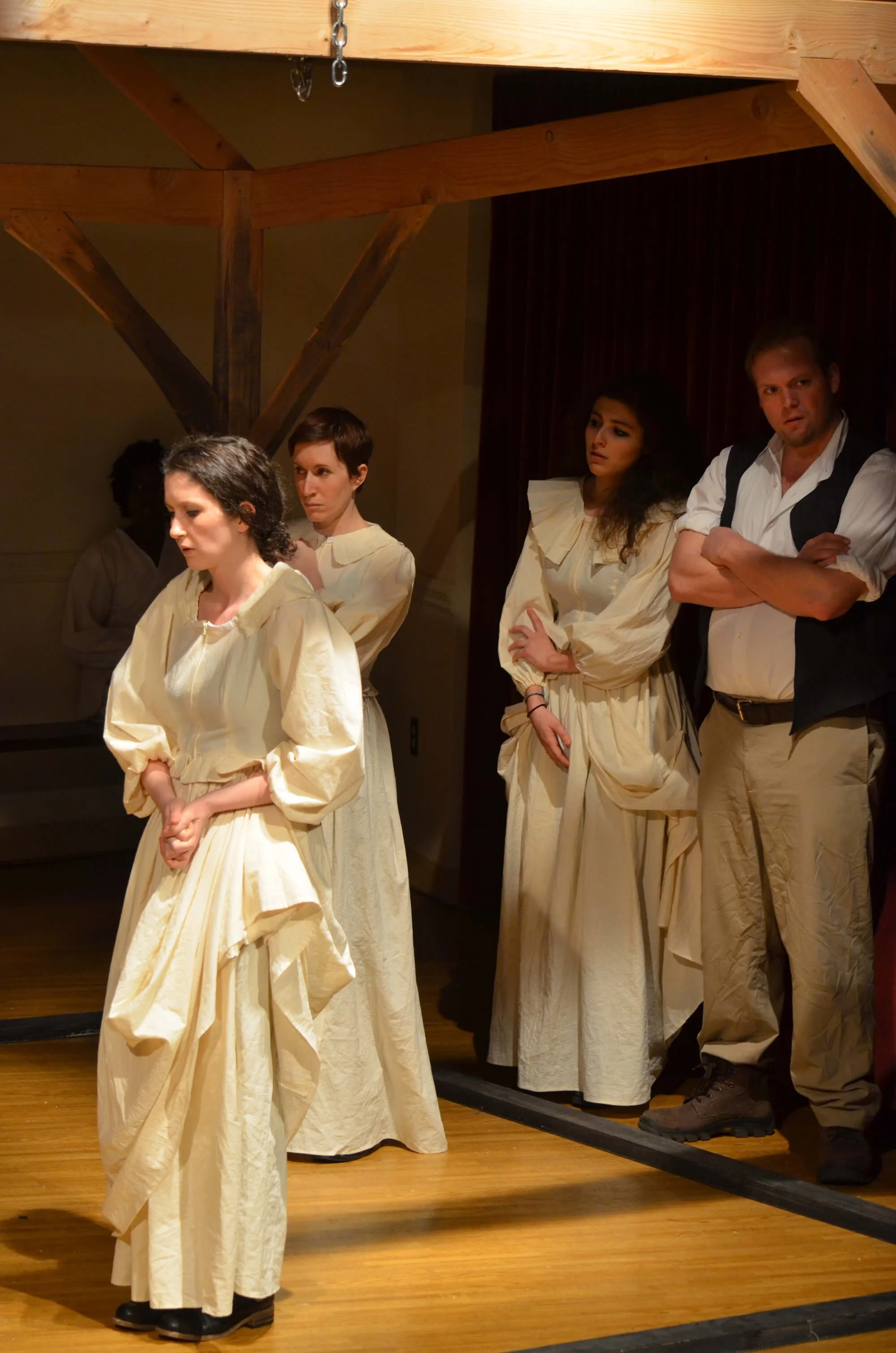 Group of four people standing on a wooden stage, dressed in historical or theatrical costumes, with serious expressions, in a theatrical setting.