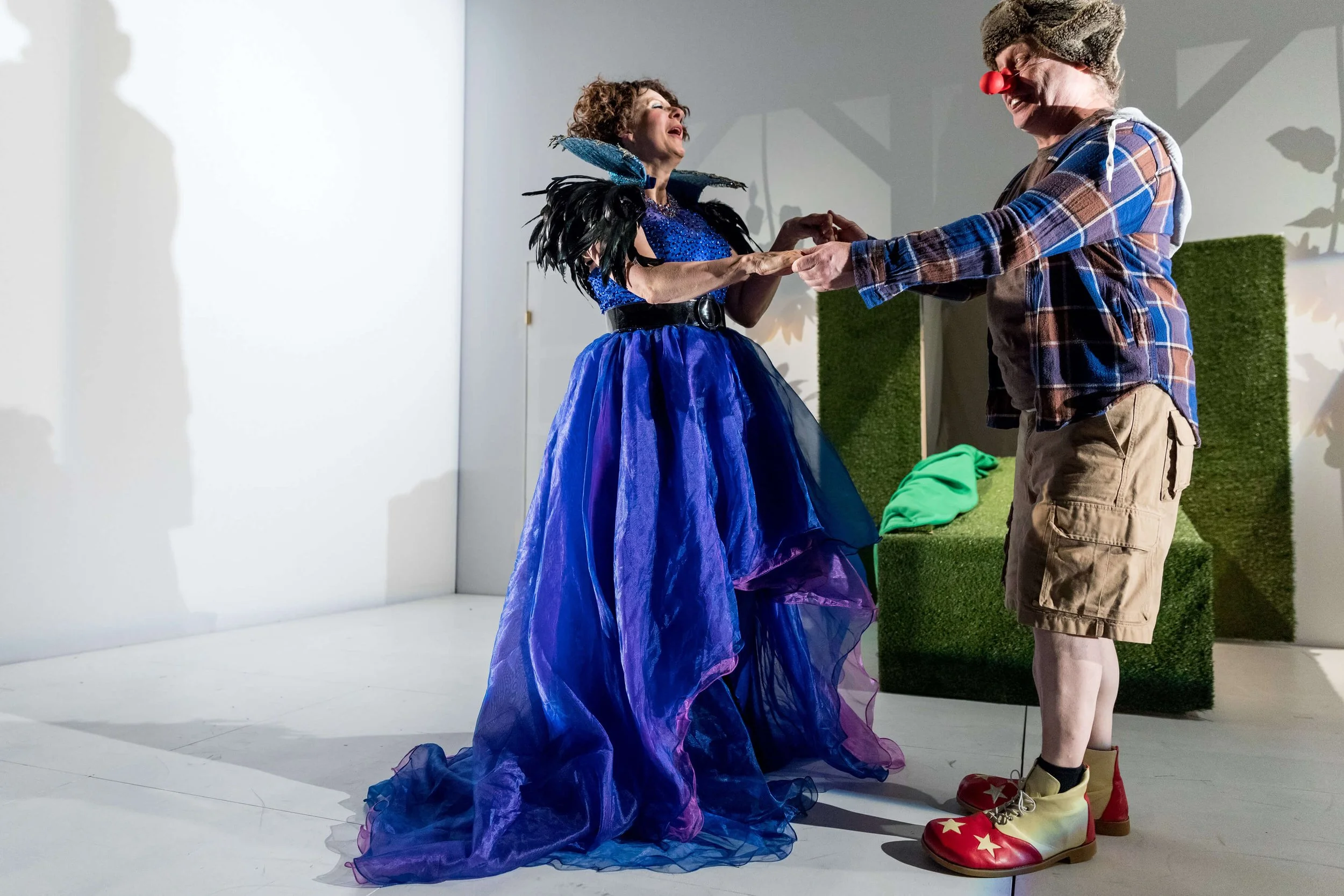 A woman in a blue dress with black feathered shoulders and a fake black belt is dancing with a man dressed as a clown. The clown has a red nose, a brown fur hat, plaid shirt, khaki shorts, and large clown shoes with stars and yellow and red colors.