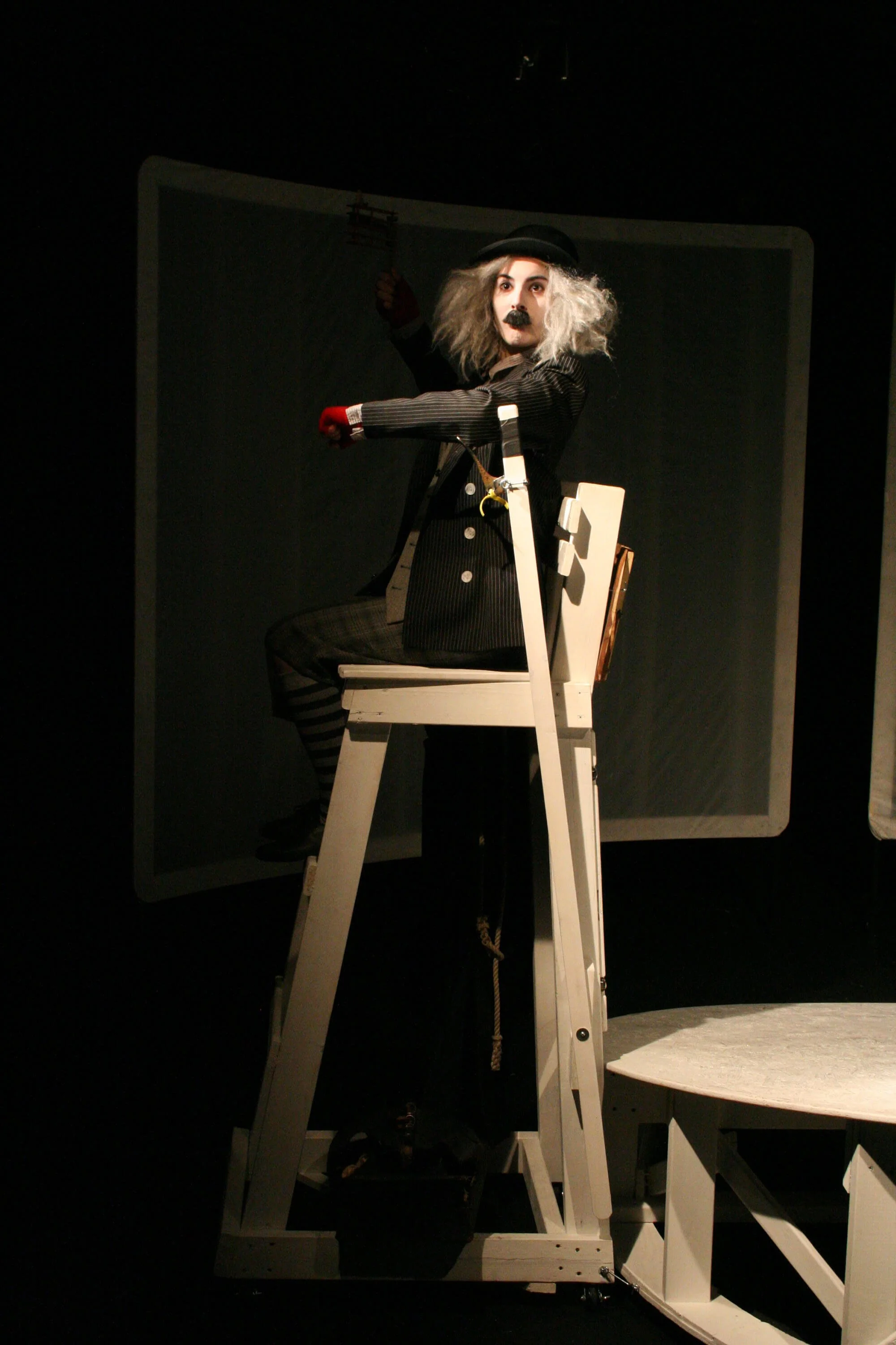 Performer dressed as a mime in a black suit with striped shirt, sitting on a white chair on a ladder, with a mirror in the background, on a dark stage.