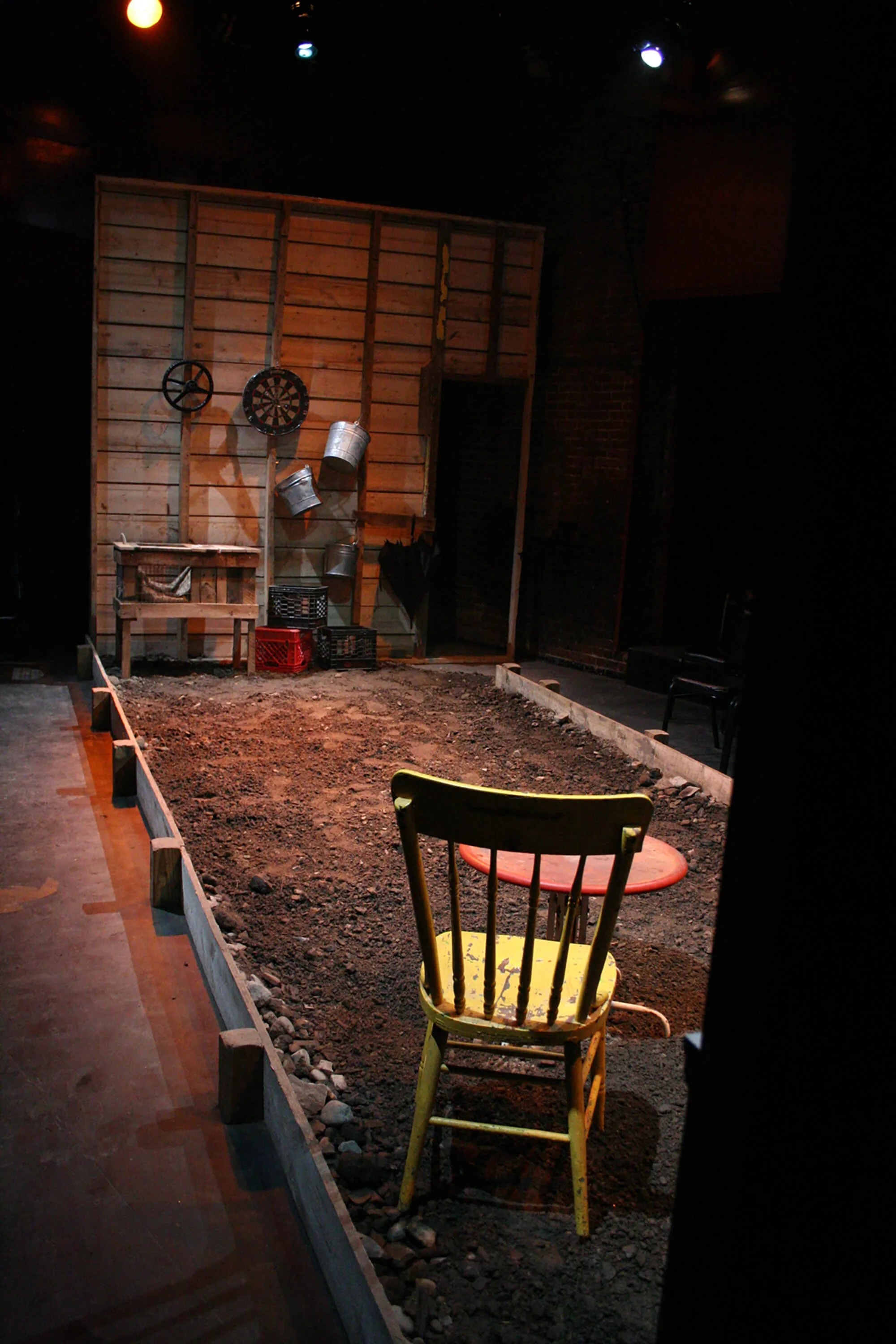 An indoor stage set resembling a rustic outdoor scene with a wooden backdrop, a yellow chair, a red plastic table, and various props like buckets, a dartboard, and a small workbench.