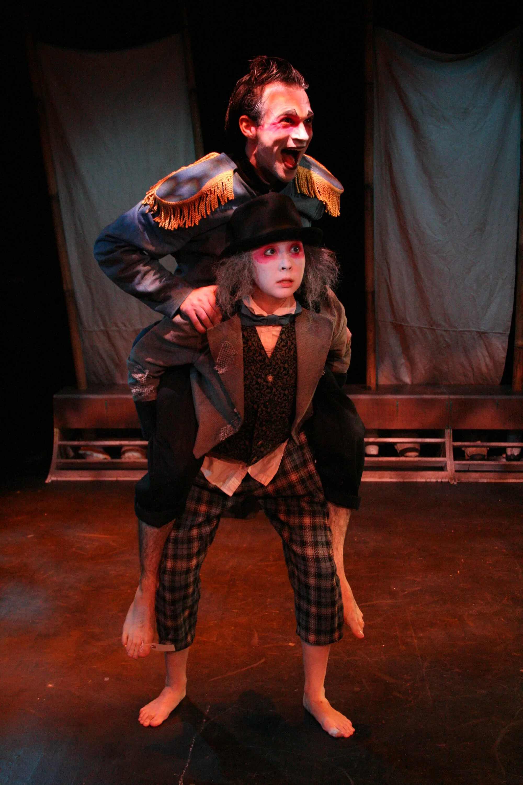 Two actors performing on stage, one standing behind the other. The actor in front is wearing a plaid pants, a jacket, a vest, a shirt, and a black hat. The actor behind is in a costume with gold epaulets, has painted face, and hair styled in a slicke