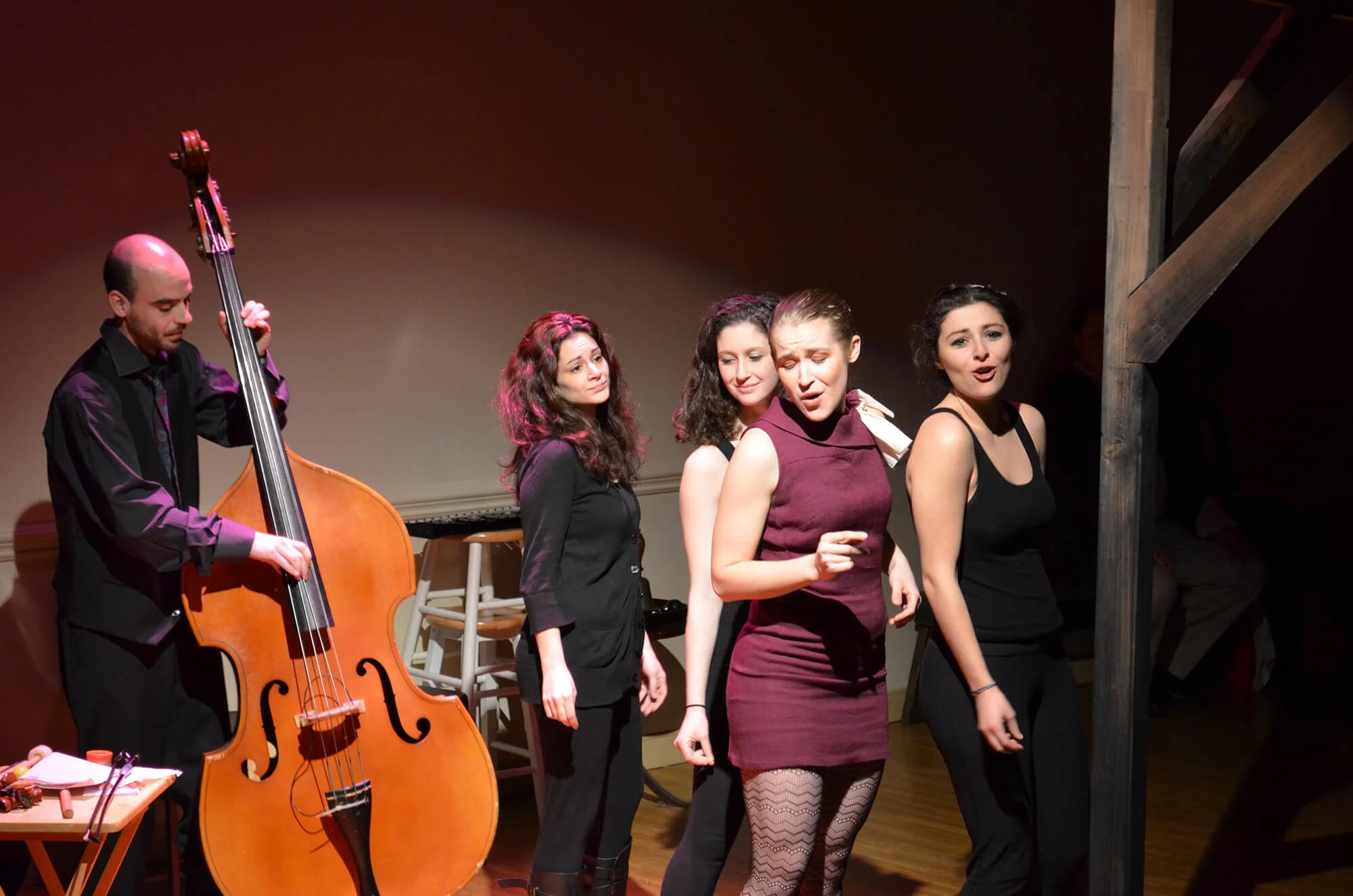 Five performers on stage, three women singing, one man playing double bass, with warm stage lighting.