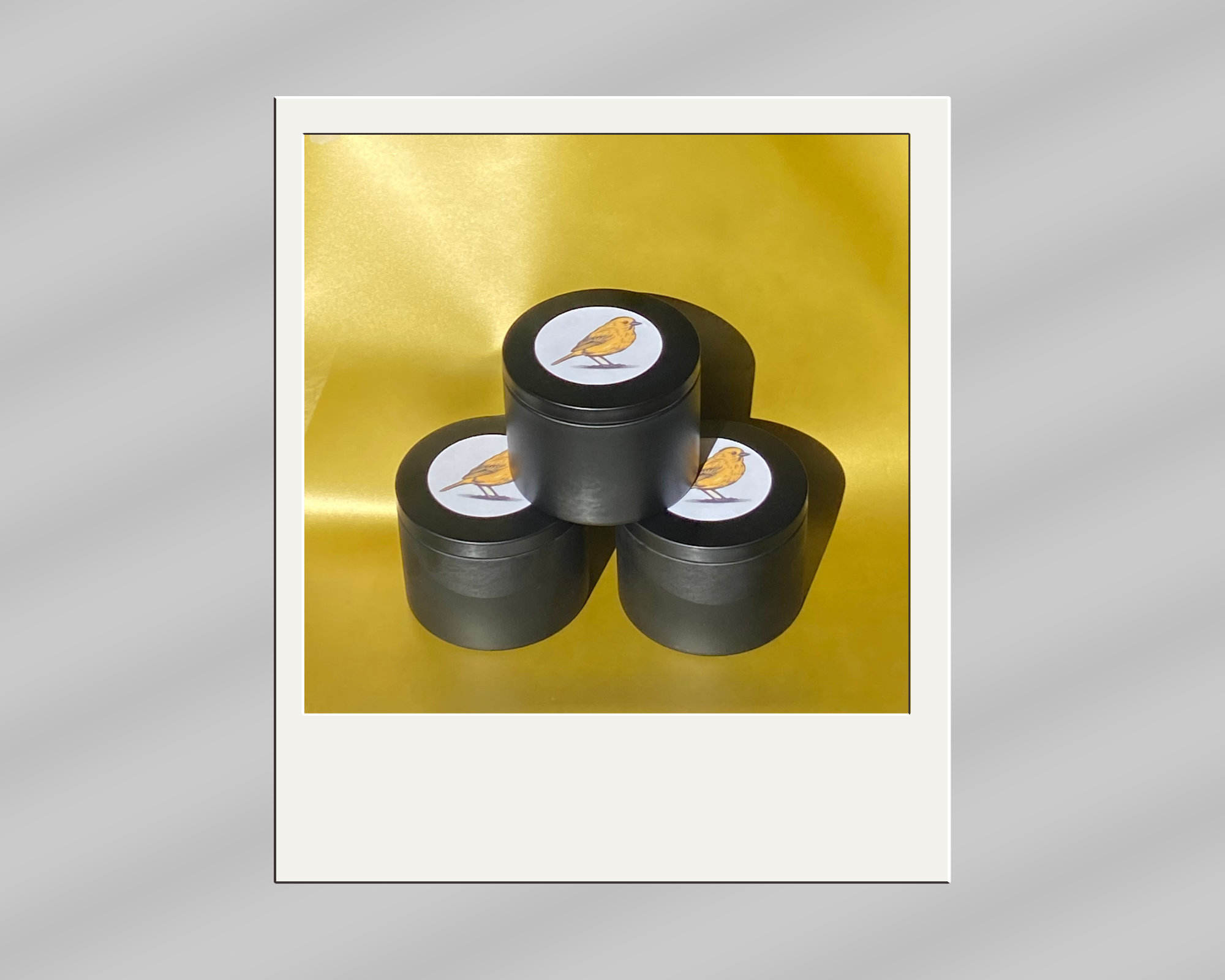 Three black containers with labels showing a yellow bird on a white background, arranged in a pyramid formation on a yellow surface with a yellow backdrop.
