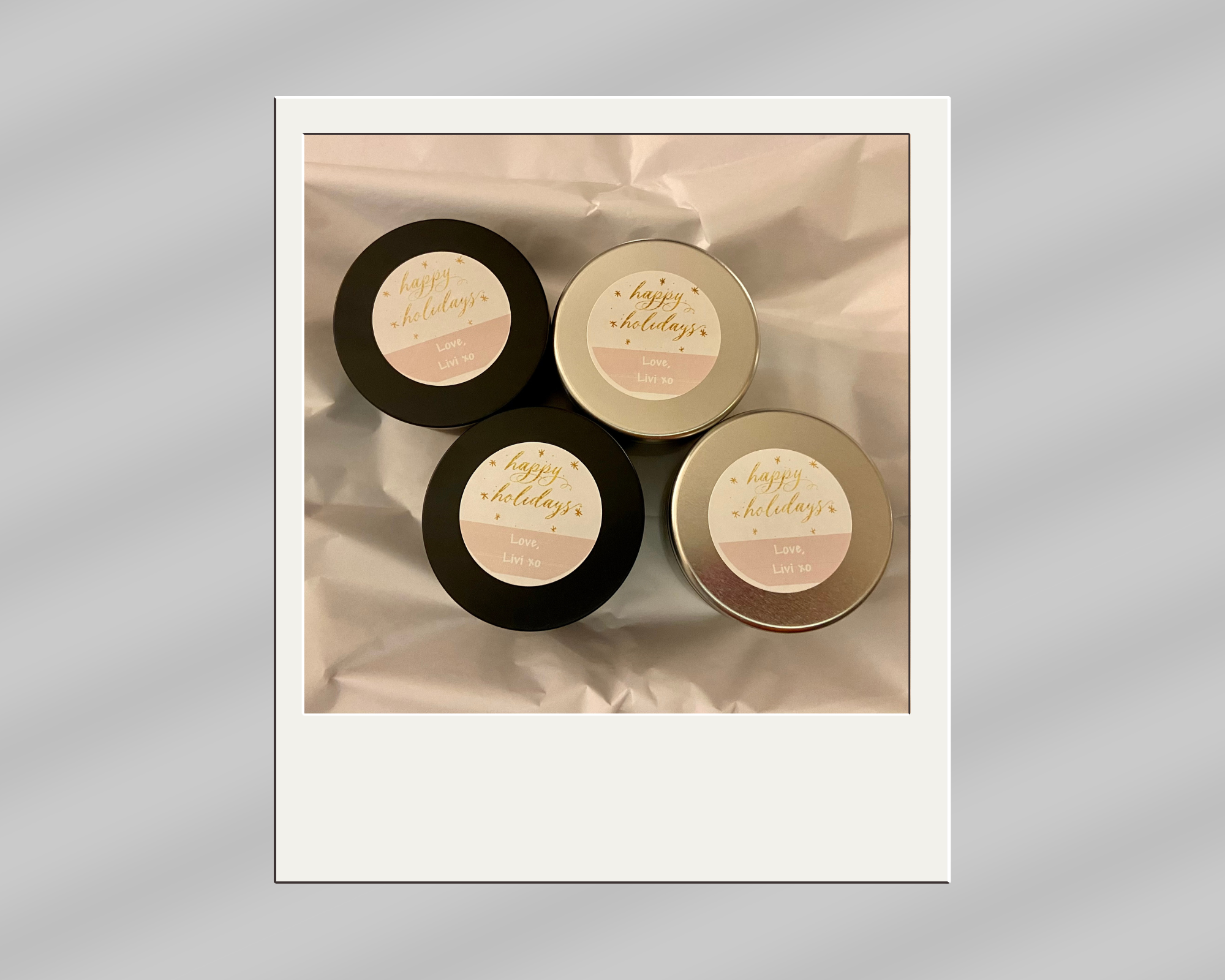 Four round tins with holiday greeting labels, two in black and two in white, arranged on crumpled white paper.