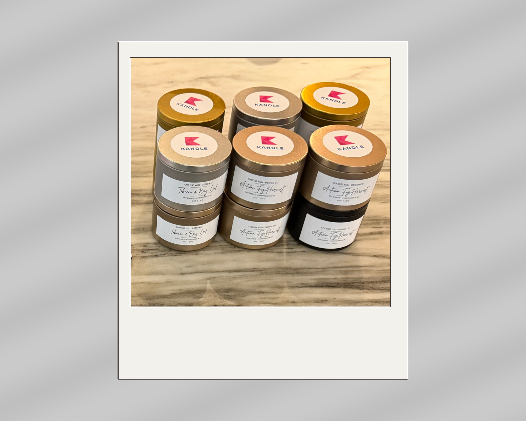 A collection of six candles in small jars arranged on a wooden surface, with two labels reading 'KANDLE' on the tops, and white labels on the sides with handwritten names of different scents.
