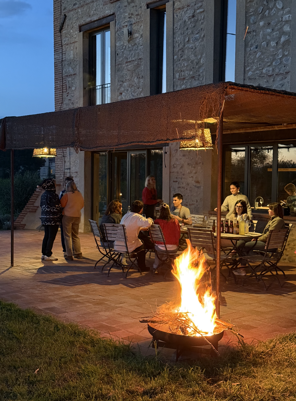 Un groupe de personnes assises et discutant autour d'une table lors d'une soirée en plein air, avec un feu de camp allumé devant eux. La scène se déroule devant une maison en pierre avec de grandes fenêtres, sous une couverture en tissu rouge-orangée