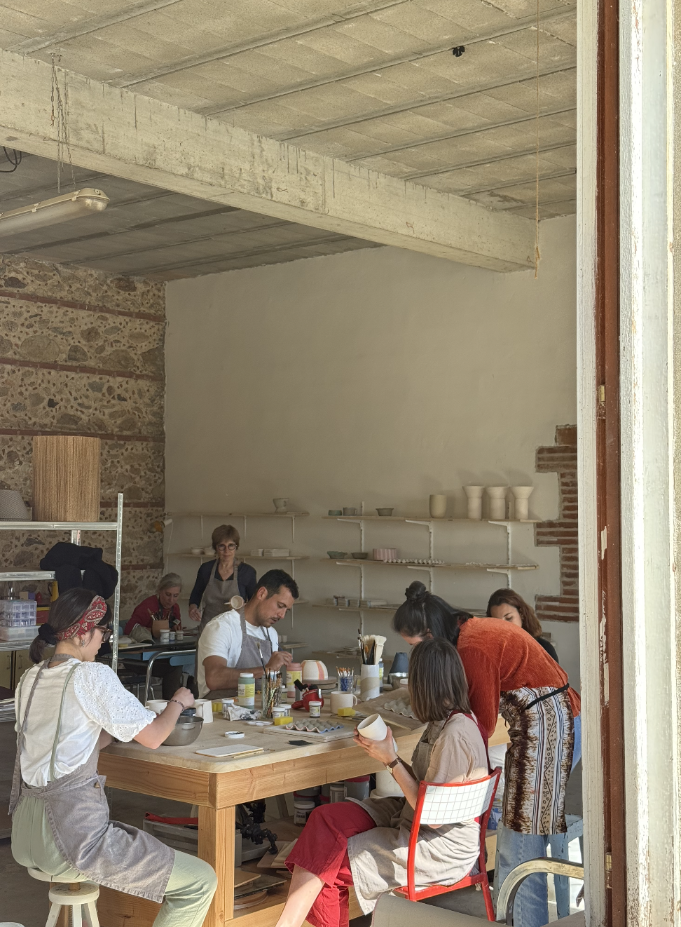 Un groupe de personnes travaillant sur des projets de poterie dans un atelier, avec des étagères de céramiques terminées en arrière-plan.