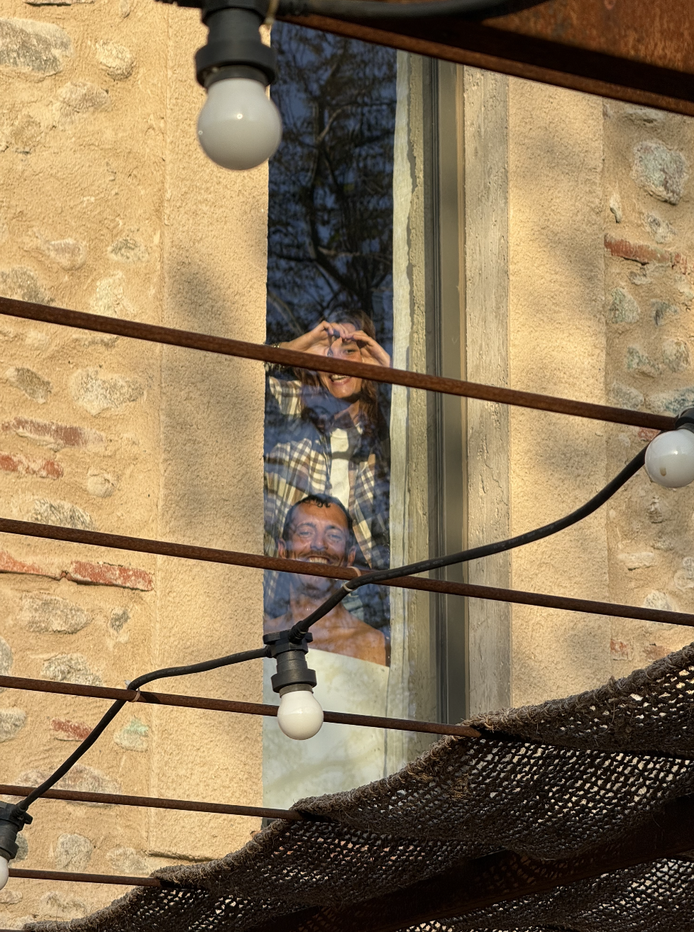 Une femme et un homme regardant par une fenêtre, leur reflet visible, dans un bâtiment en pierre, éclairés par des ampoules suspendues.