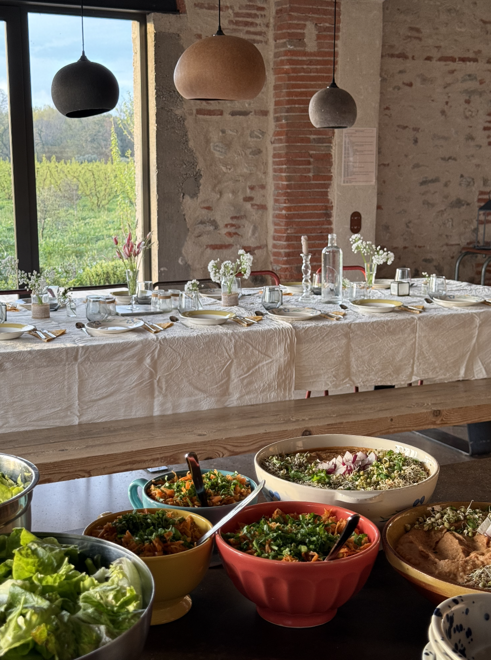 Table dressée pour un repas avec des plats à saladier, une salle lumineuse avec des lampes suspendues et une grande fenêtre laissant apparaître un paysage verdoyant.