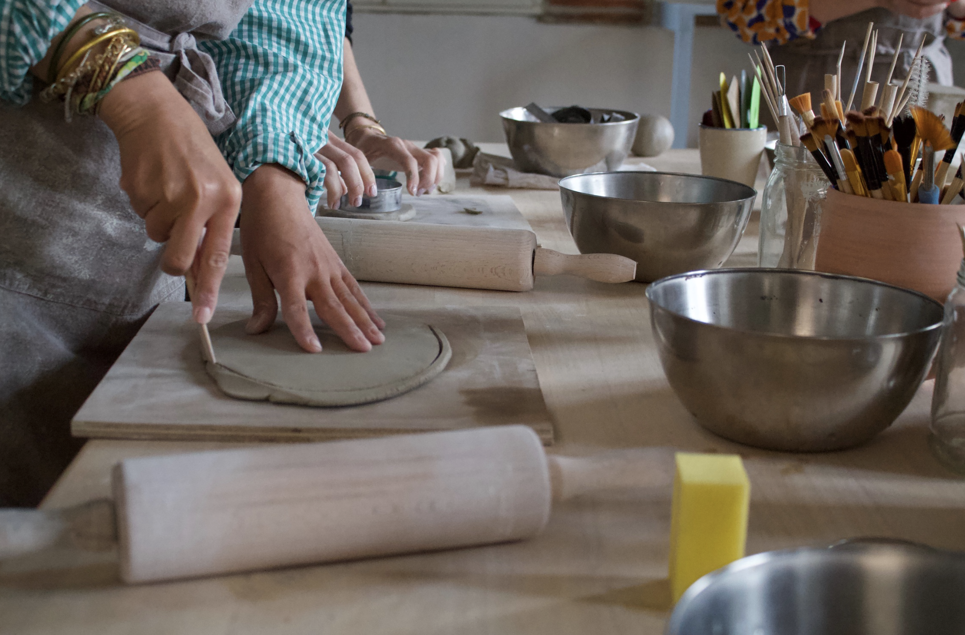 Groupe de personnes utilisant un rouleau à pâtisserie pour étaler de la terre sur une surface en bois, entouré de divers ustensiles de céramique.