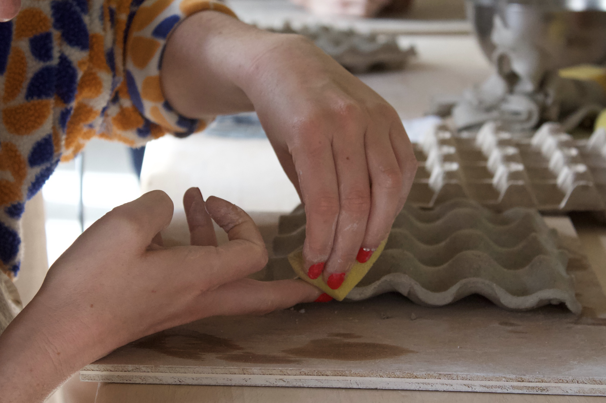 Deux mains, dont une avec des ongles rouge, manipulent une pâte ou une boule dans un moule en carton ondulé sur une surface en bois, avec des objets flous en arrière-plan.