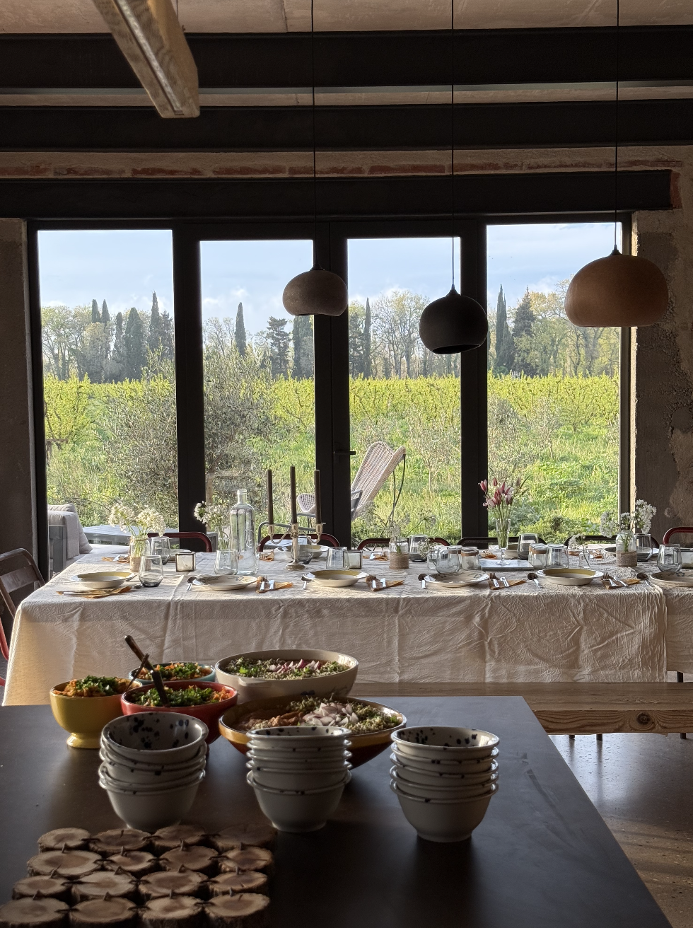 Une salle à manger avec une grande table recouverte d'une nappe blanche, dressée pour un repas, avec des assiettes, des verres, des bougeoirs et des fleurs. En arrière-plan, une grande fenêtre donnant sur un paysage de champ avec des arbres et un cie