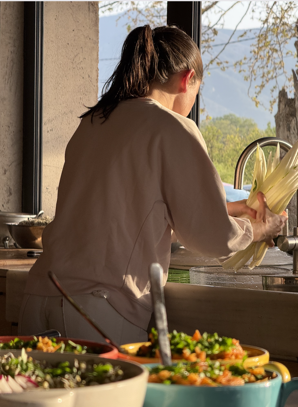 Une femme prépare des légumes dans une cuisine près d'une fenêtre avec vue sur la nature.