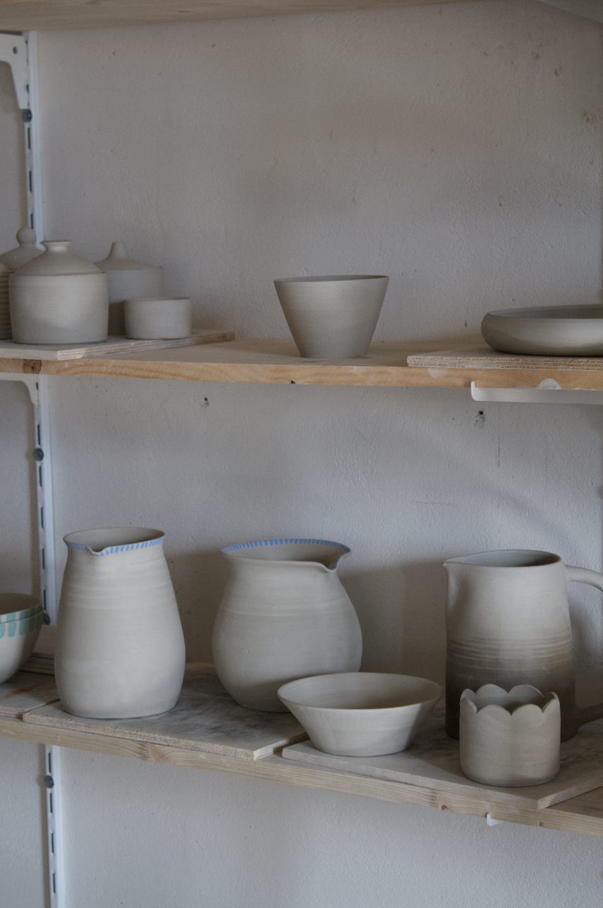 Étageres avec des poteries en céramique non émaillées, incluant des vases, bols, et un pichet, dans un atelier ou une boutique de poterie.
