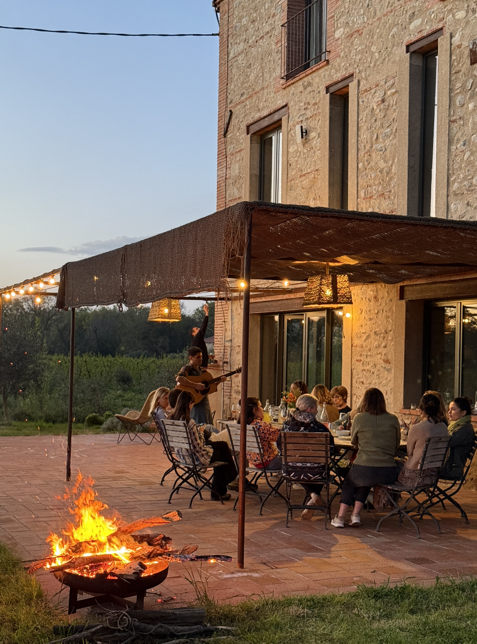 Groupe de personnes dégustant un dîner en plein air sous un auvent, avec un musicien jouant de la guitare, devant une maison en pierre, en soirée, avec un feu de camp en premier plan.