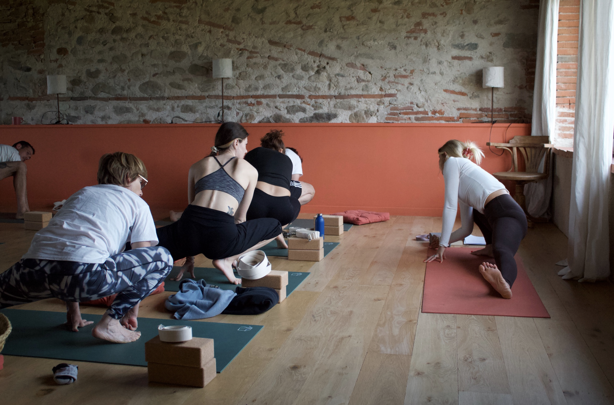 Groupe de personnes suivant un cours de yoga dans une salle avec mur en pierre et sol en bois, une personne pratique la posture sur un tapis rouge.