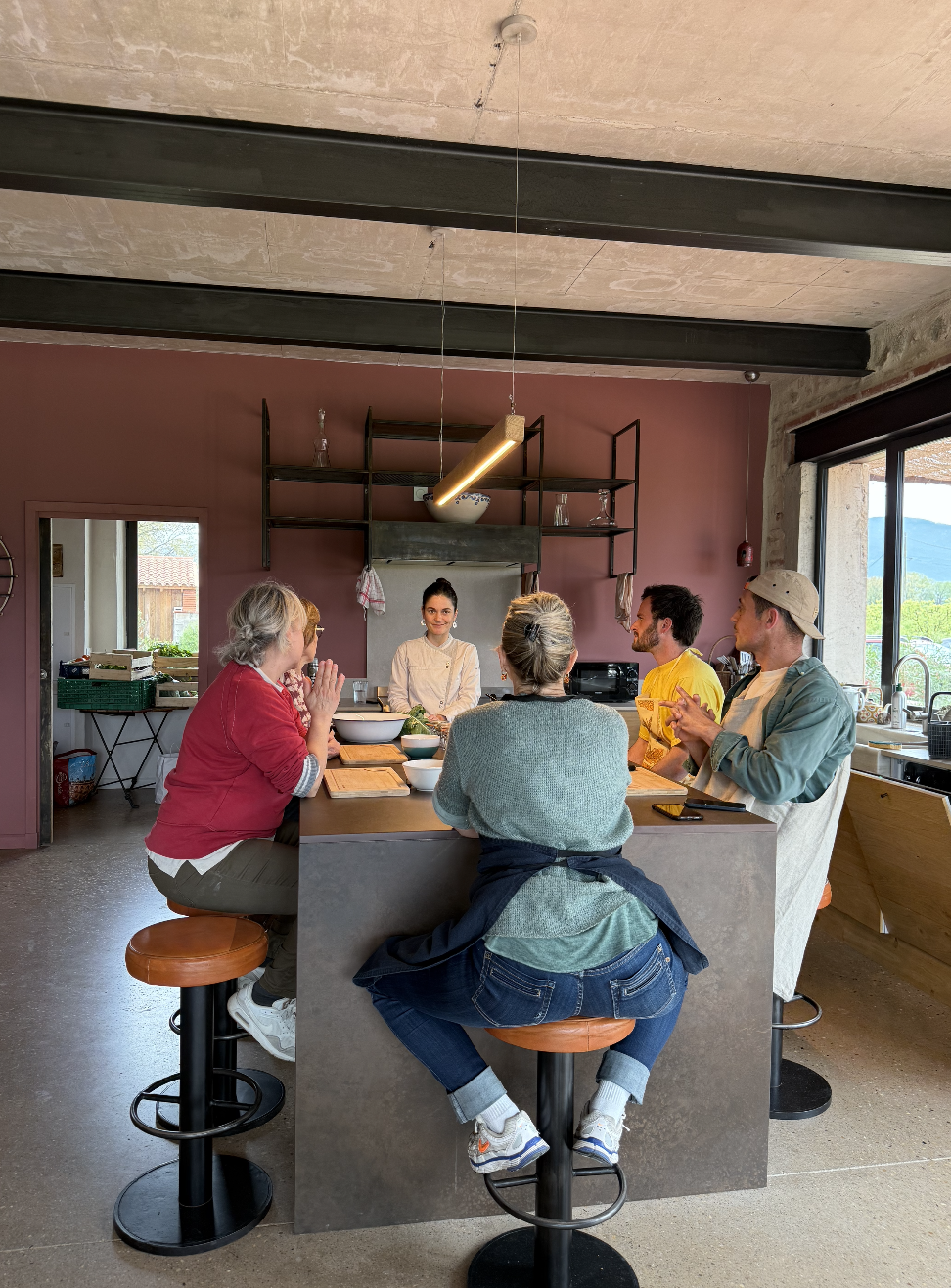 Groupe de six personnes, dont une femme qui semble être une chef, assises autour d'un îlot de cuisine dans une pièce moderne, en train de discuter et d'écouterla chef.