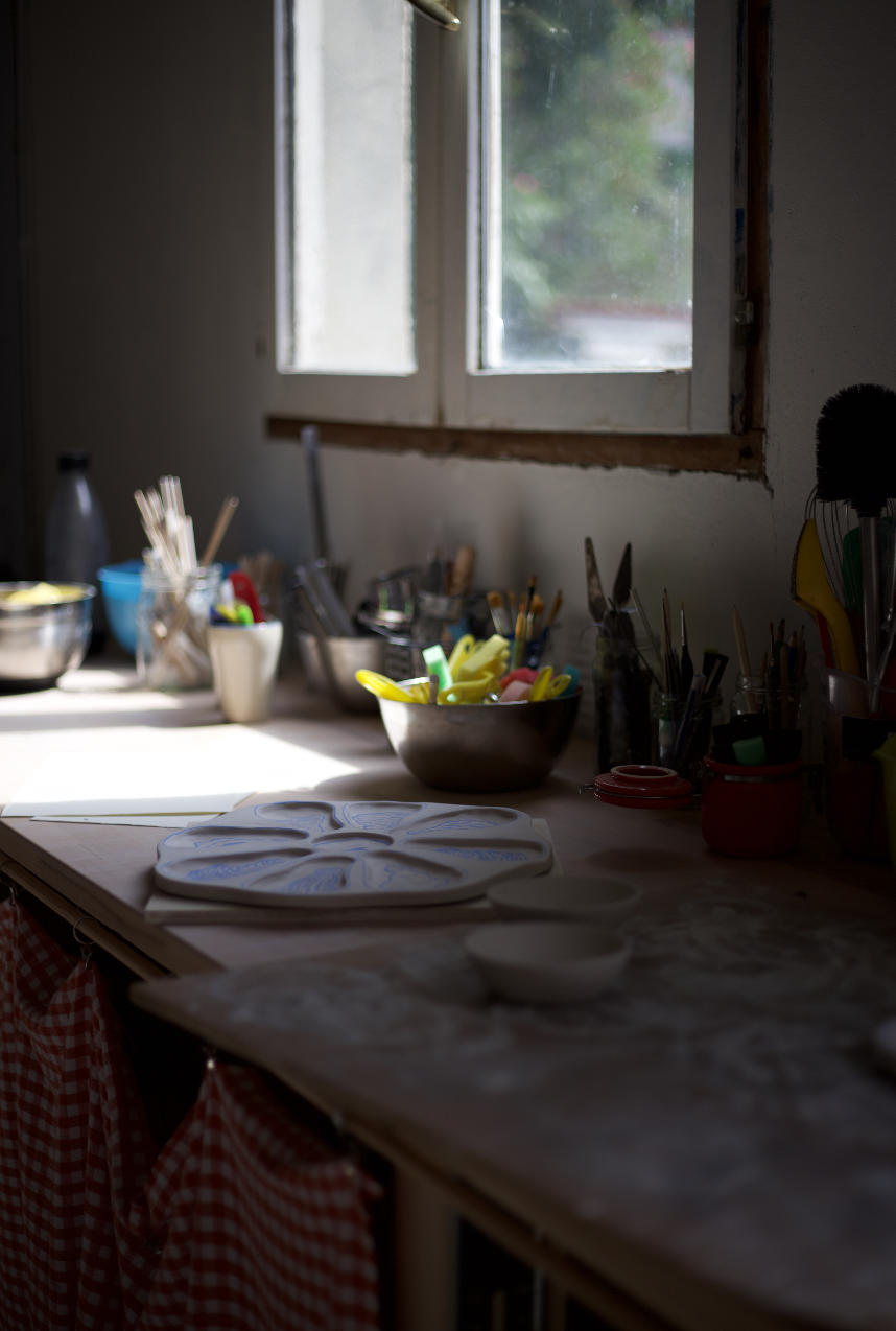 Une cuisine ou un atelier de pâtisserie avec des ustensiles, des bowls, et des plans de travail en partie éclairés par la lumière naturelle passant par une fenêtre
