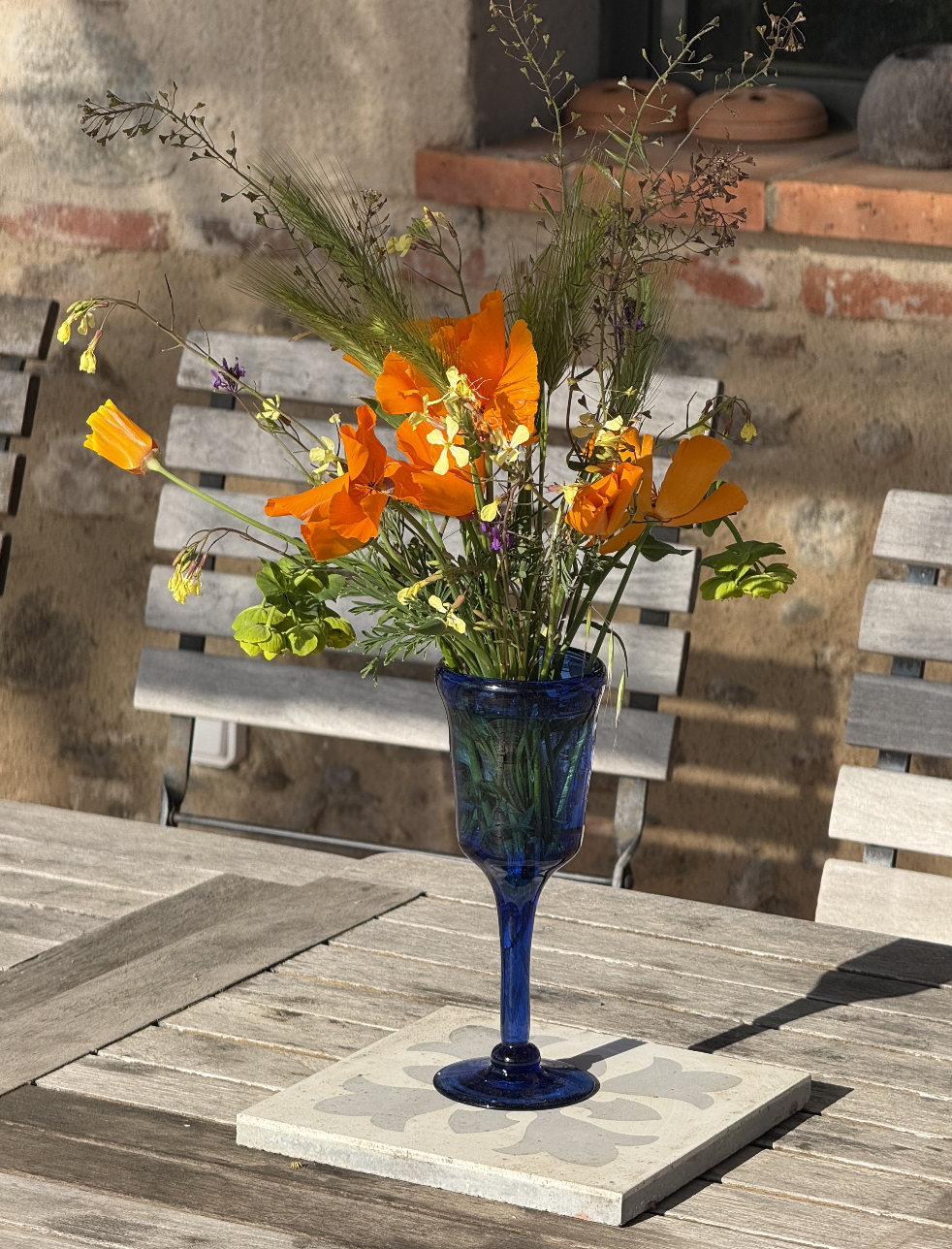 Un bouquet de fleurs colorées dans un vase en verre bleu, placé sur une table en bois, avec des chaises en arrière-plan et un mur en pierre et maçonnerie derrière.