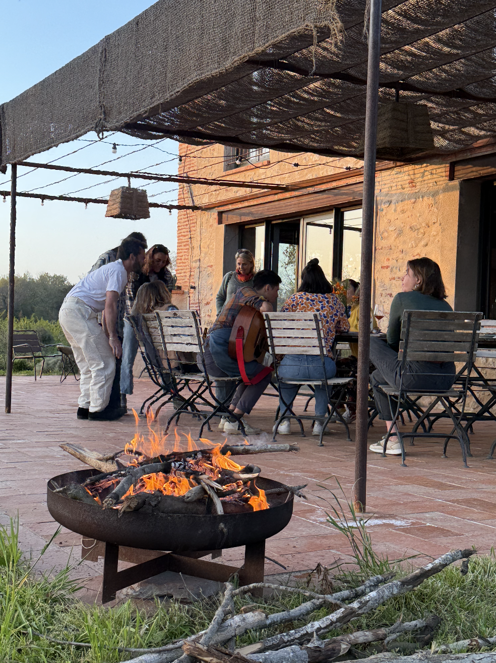 Un groupe de personnes assises autour d'une table de restaurant en extérieur, avec un feu de bois brûlant au premier plan, sous une structure de toile.