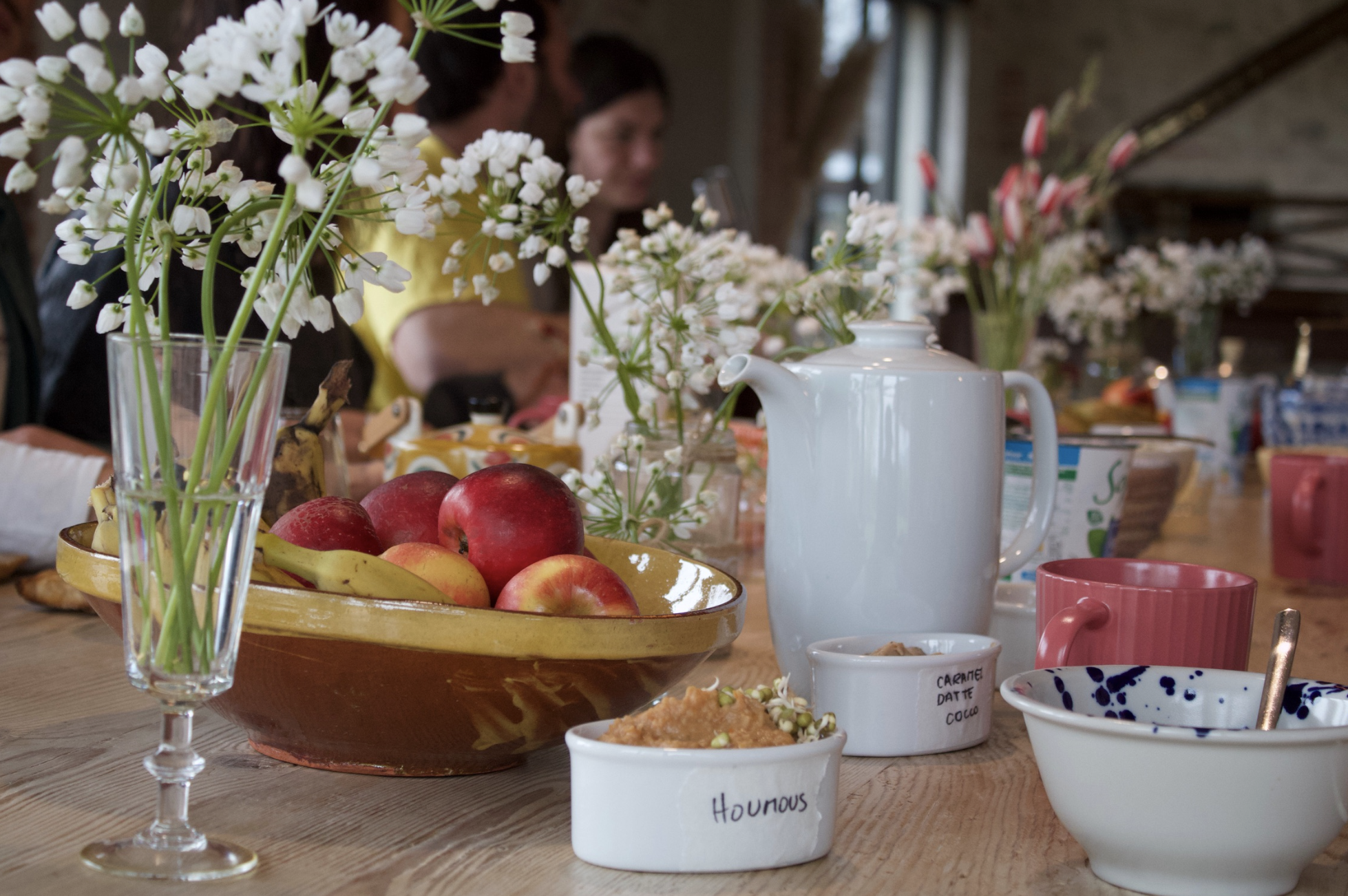 Table dressée avec des fleurs, pommes, pain, yaourt, confitures, tasses, et une théière, dans une ambiance de petit-déjeuner