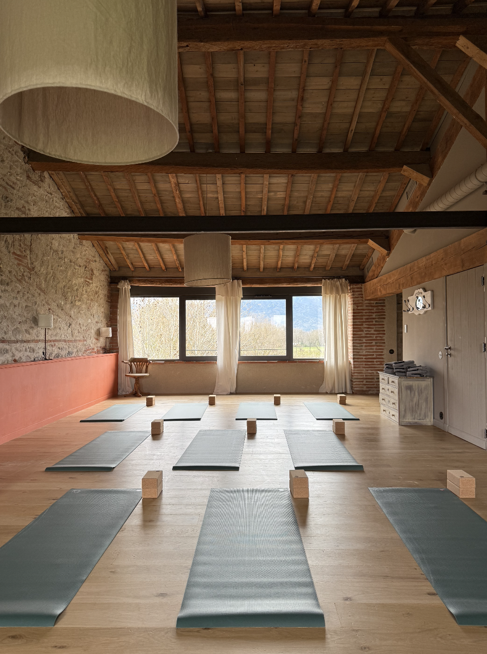 Salle de yoga avec des tapis et des blocs en bois, grande fenetre avec vue sur la nature, murs en pierre et plafond en bois exposé.