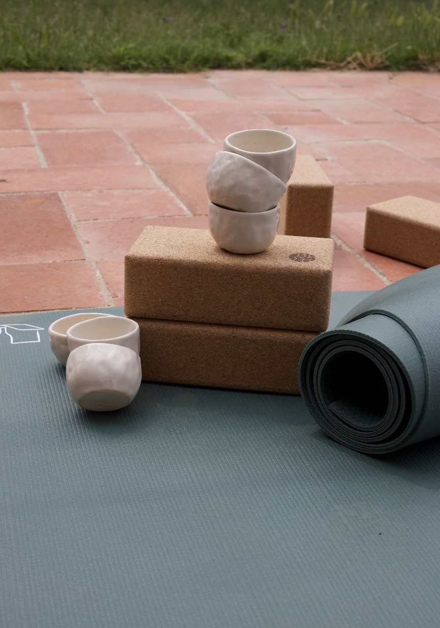 Tasse en céramique posée sur des blocs de liège, accompagnée d'autres tasses, un tapis de yoga roulé et un tapis de yoga bleu, avec un sol en tuiles en arrière-plan.