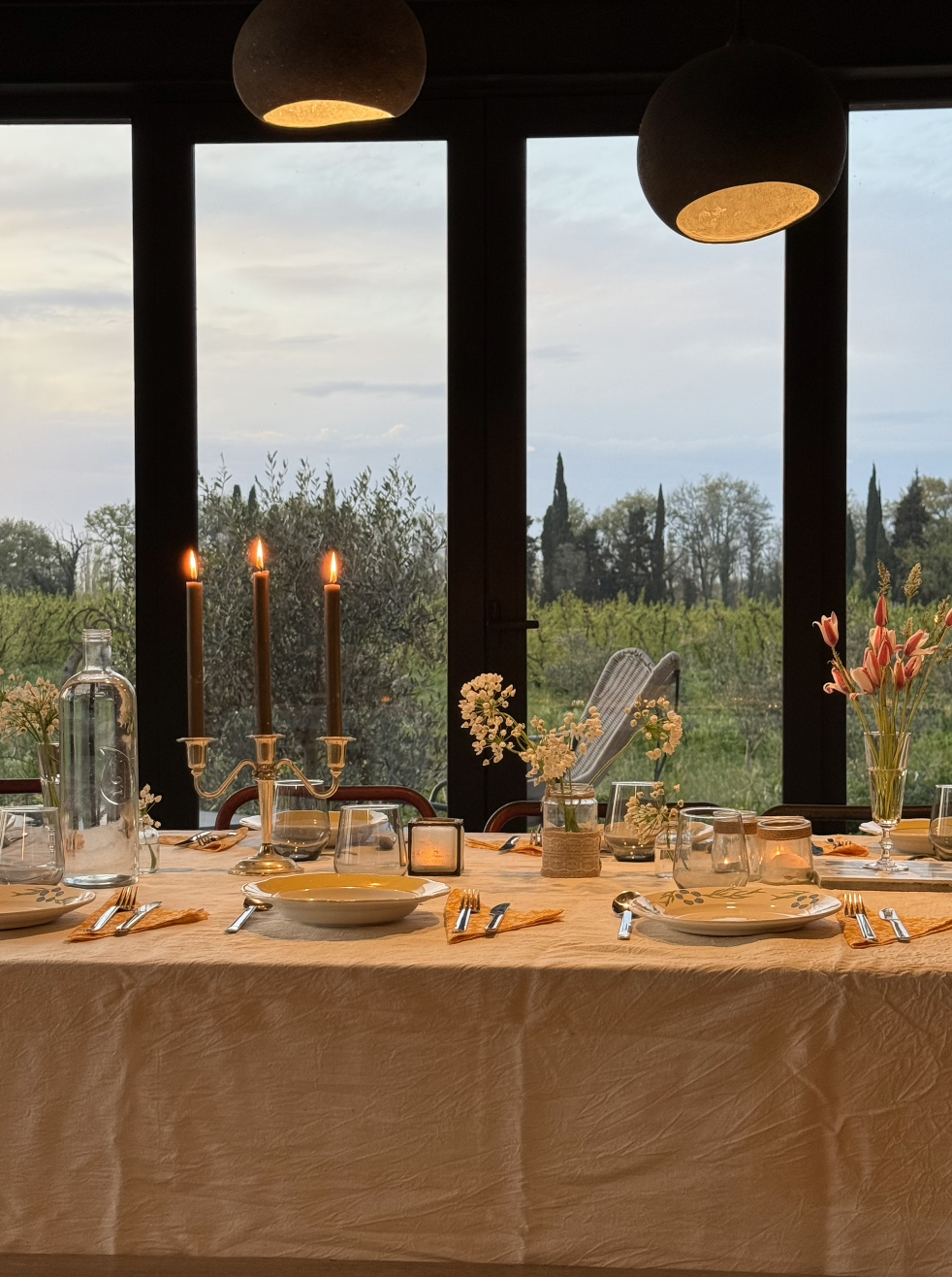 Une table élégante avec une nappe beige, décorée de fleurs, de bougies, de vaisselle, de fourchettes, de cuillères et de verres. En arrière-plan, une vue sur un paysage verdoyant avec des arbres, à travers de grandes fenêtres, sous un ciel légèrement