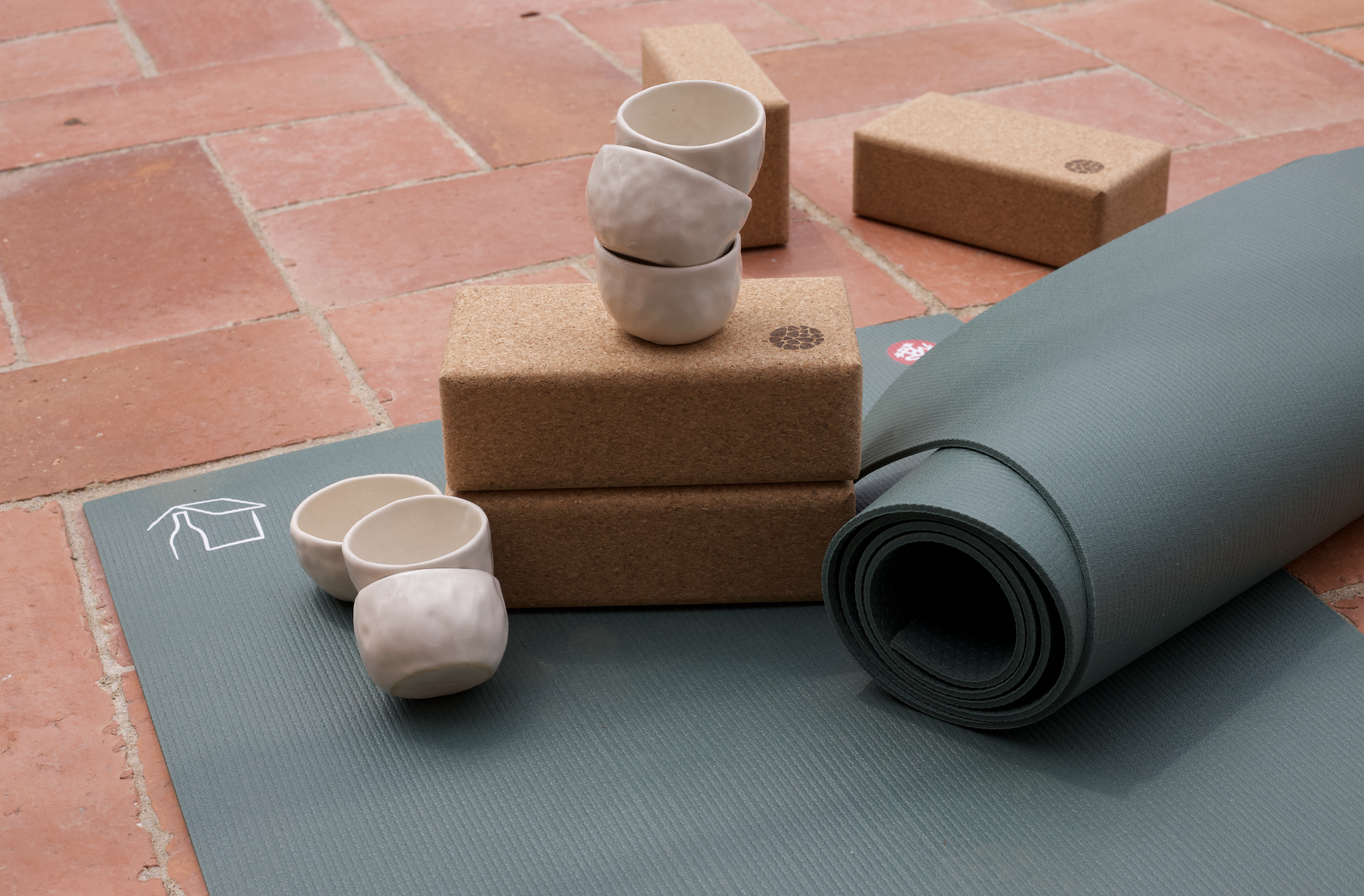 Un tapis de yoga gris roulé, deux petits bols en céramique blanche, trois tasses en céramique blanche empilées, deux briques en liège, placés sur un sol en briques rouges.