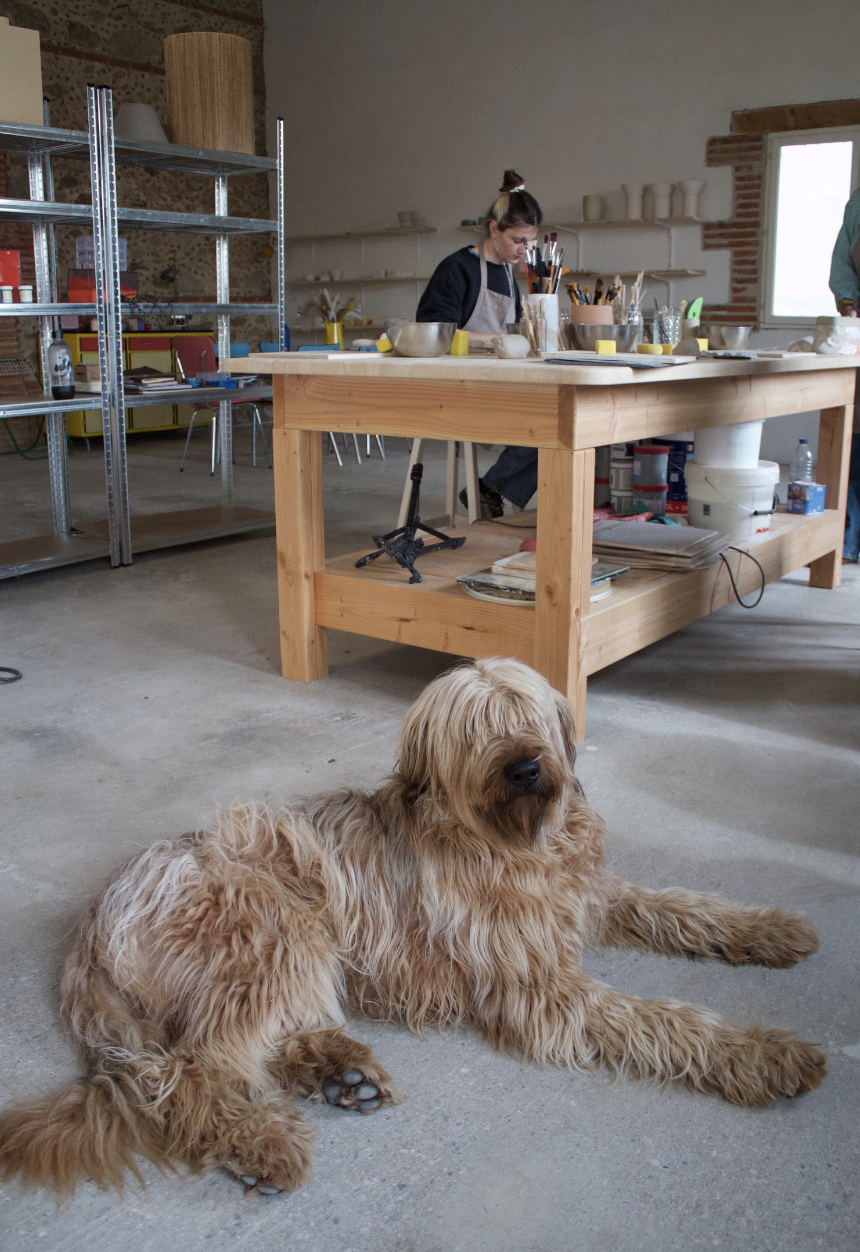 Un chien de race Goldendoodle allongé sur le sol dans un atelier tandis qu'une femme travaille à une table en bois avec des outils et des contenants.