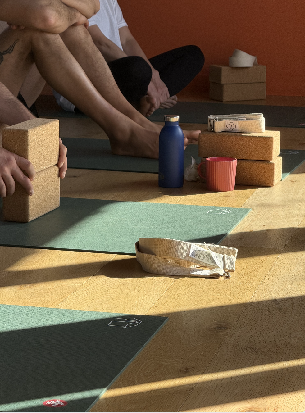 Groupe de personnes pratiquant le yoga avec tapis, blocs, bouteilles d'eau, et accessoires sur un sol en bois dans une pièce aux murs orange.