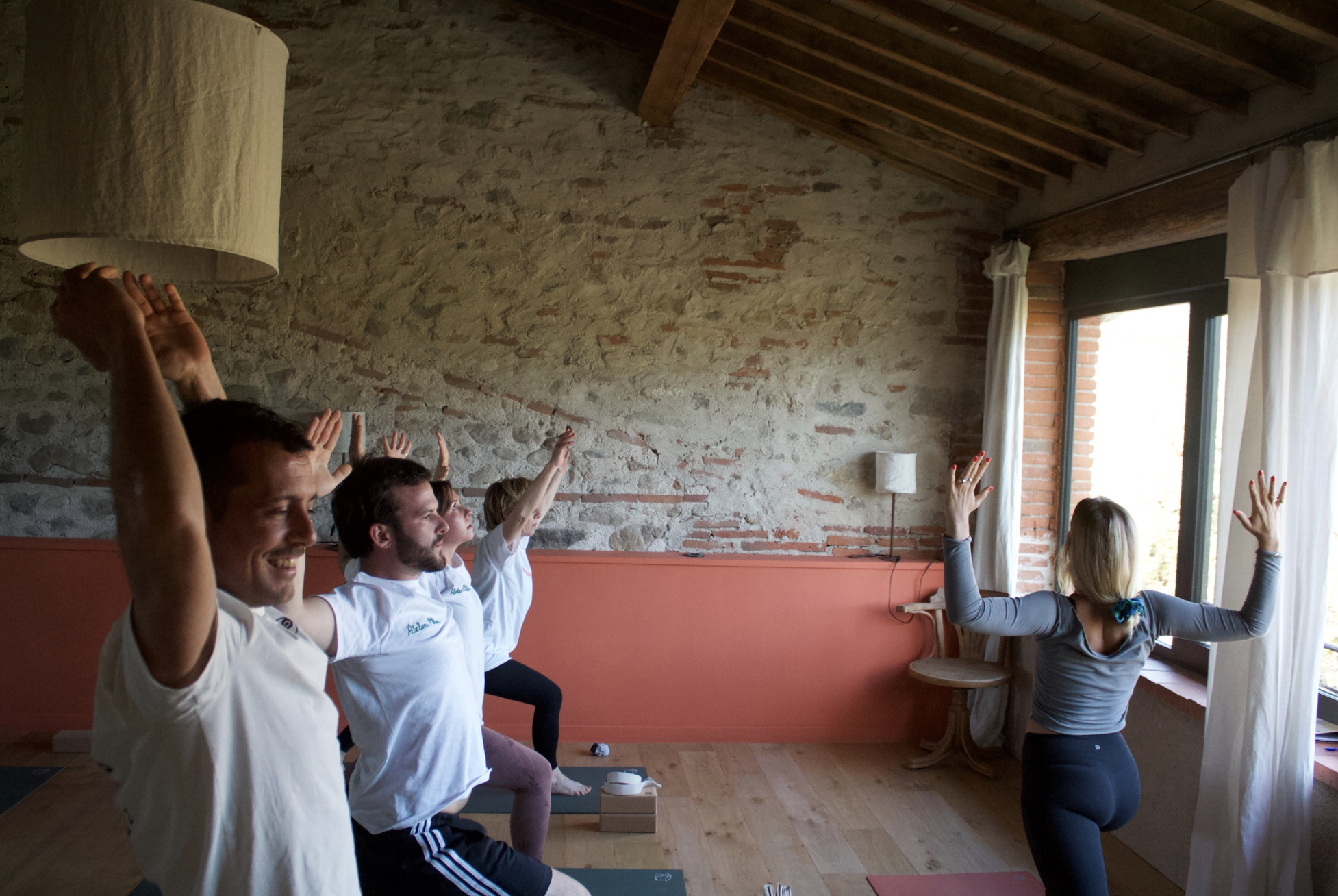 Groupe de personnes faisant du yoga dans une pièce avec murs en pierre et cheminée, en position de prière avec bras levés vers le plafond, face à une fenêtre ouverte.