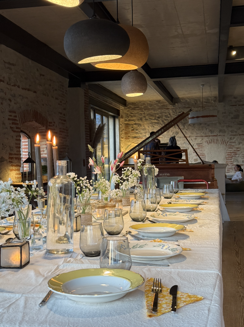 Table de dîner élégante décorée avec des fleurs, des bougies et de la vaisselle en porcelaine, dans une salle avec murs en pierre et un piano à queue en arrière-plan.